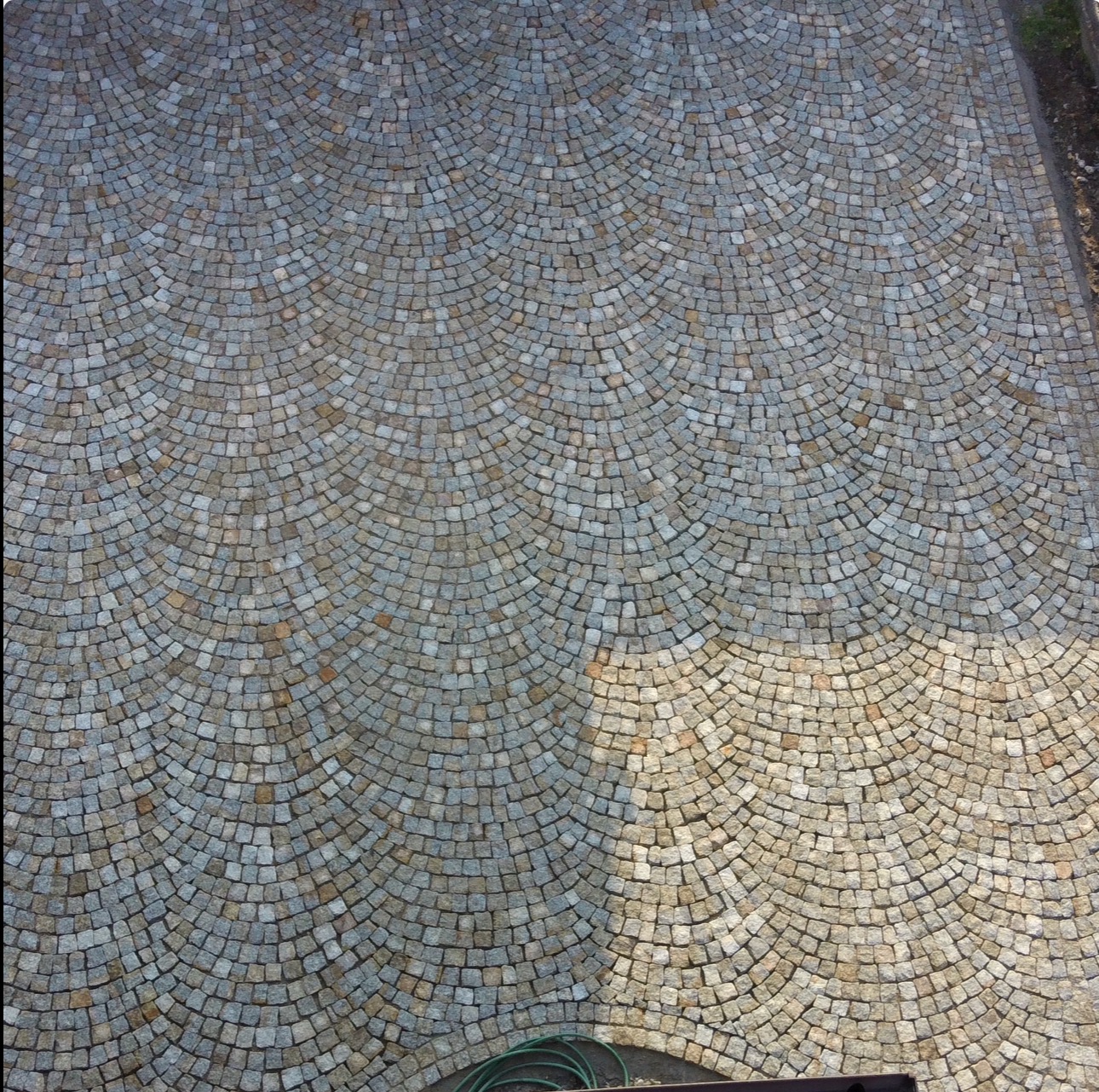 Powierzchnia wyłożona szarą kostką w łukowaty wzór, częściowo oświetlona słońcem, widok z góry.