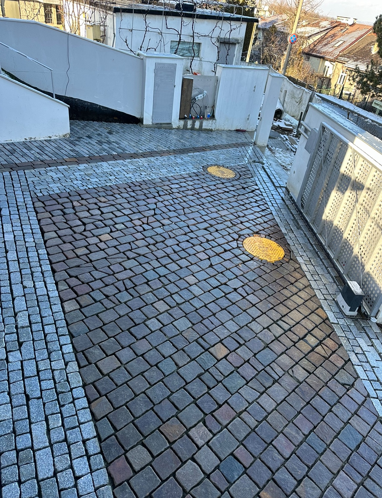 Ułożona nawierzchnia z kostki granitowej w odcieniach szarości i brązu, obramowana jaśniejszą kostką, z widocznymi studzienkami kanalizacyjnymi i fragmentem bramy wjazdowej.