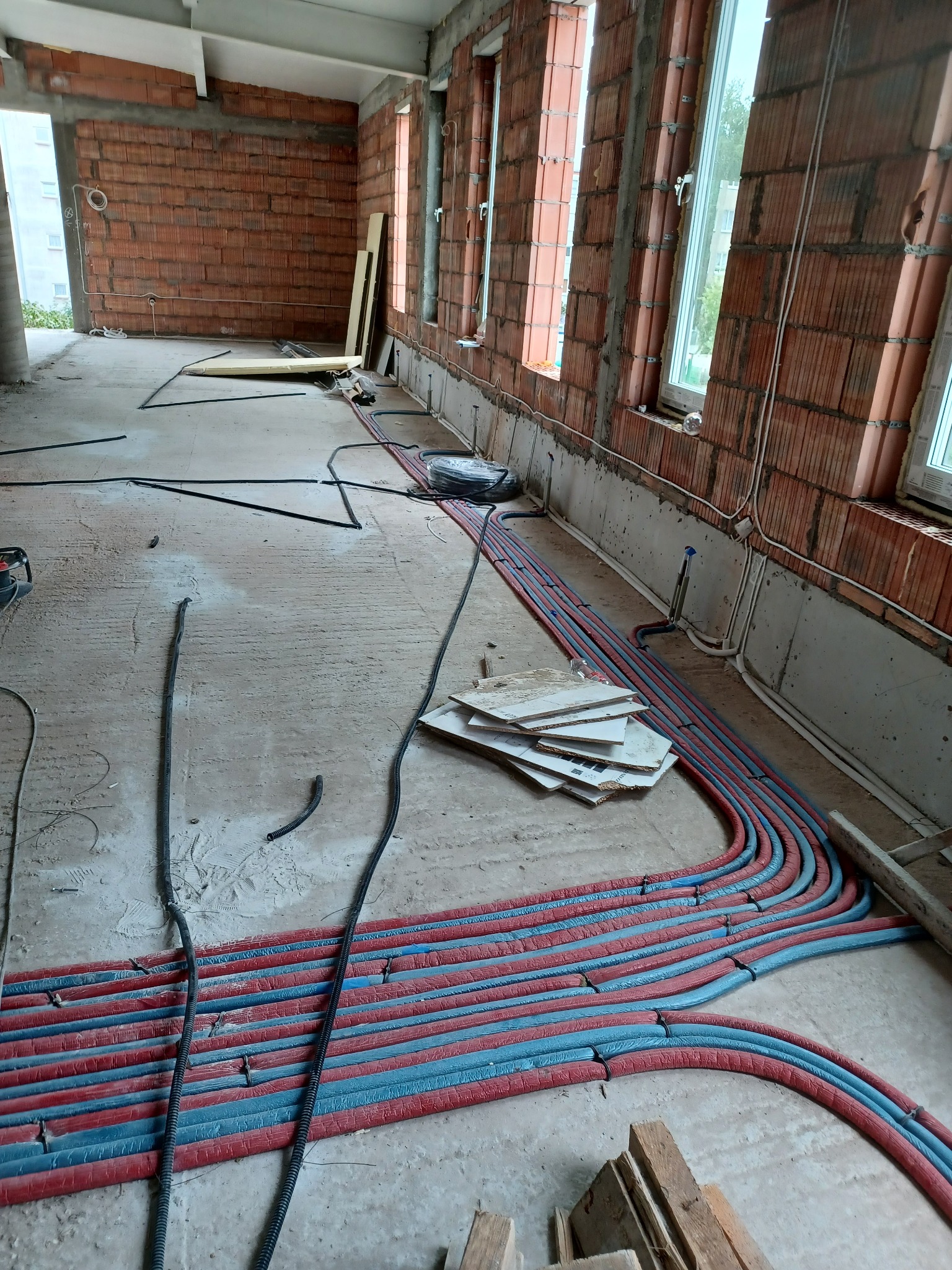 Wnętrze w trakcie budowy z widocznymi rurami grzewczymi w izolacji czerwono-niebieskiej, ułożonymi na podłodze wzdłuż ściany z oknami, z fragmentami cegieł i przewodami elektrycznymi.