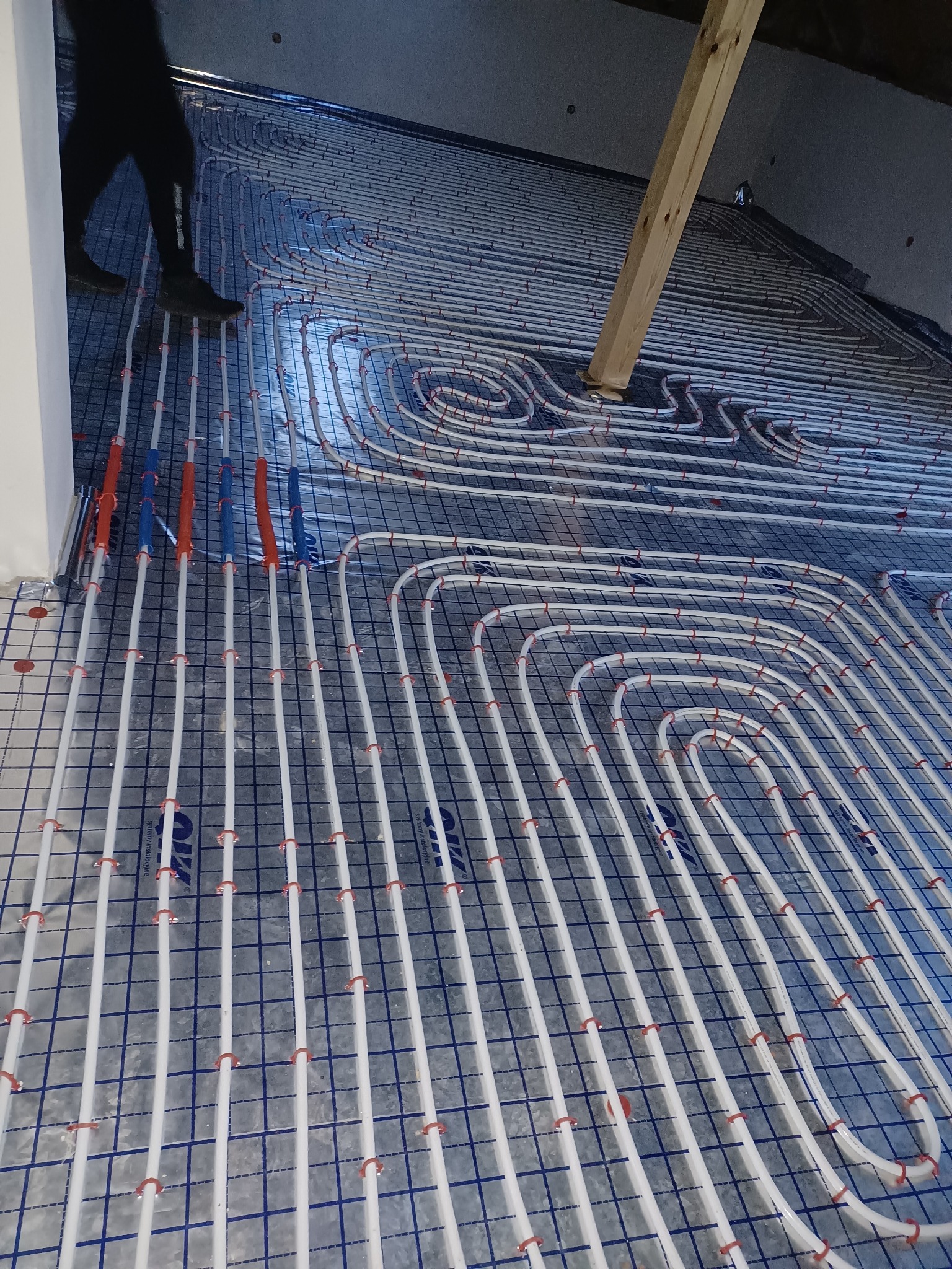 Rozłożone rury ogrzewania podłogowego na folii z rastrem, widoczne nogi instalatora w rogu kadru oraz drewniana belka stropowa