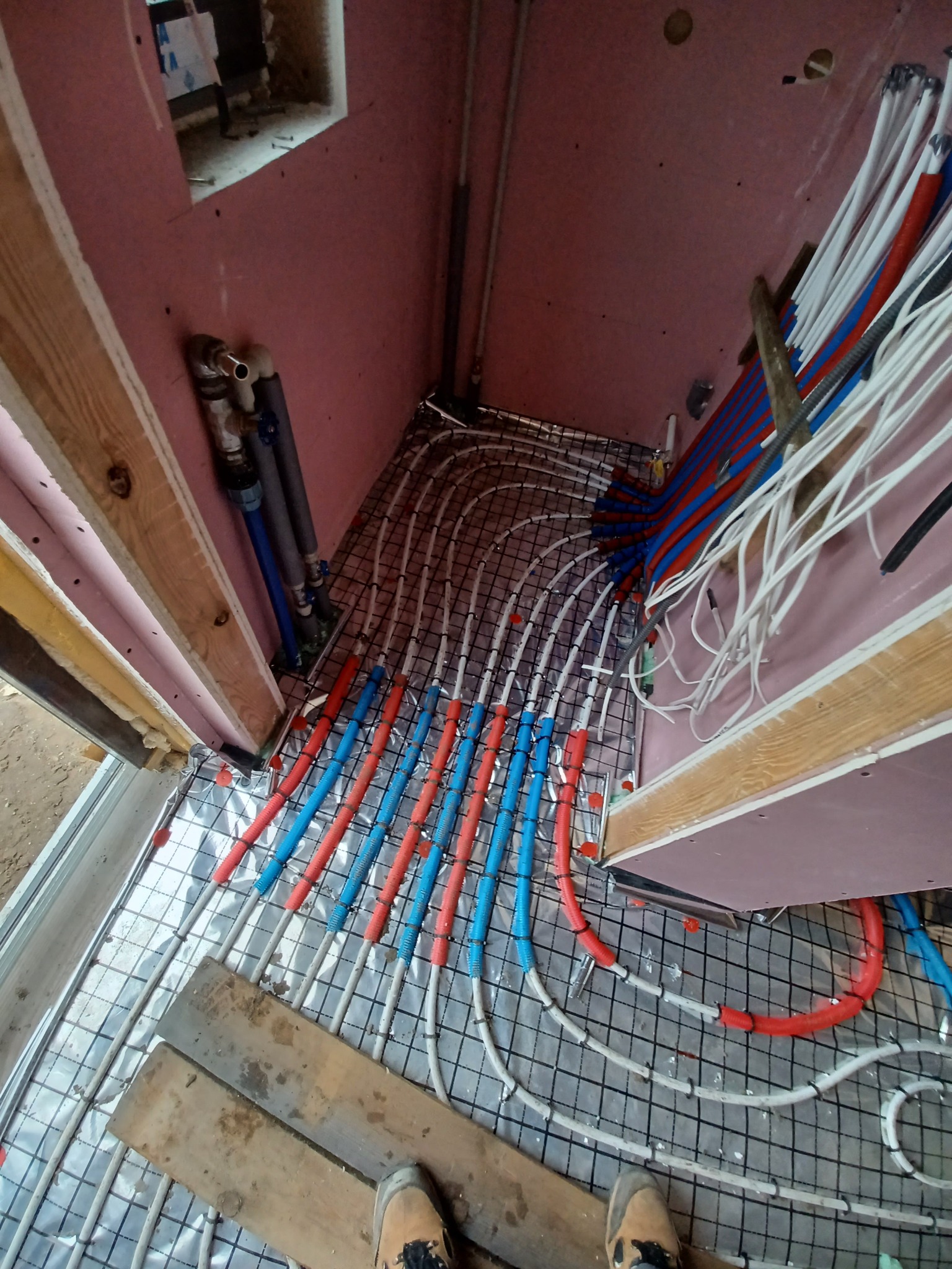 Instalacja ogrzewania podłogowego w nowo budowanym pomieszczeniu z widocznymi rurami grzewczymi w kolorach czerwonym i niebieskim, przymocowanymi do siatki na folii izolacyjnej, oraz fragmentem...