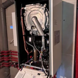 Otwarty piec gazowy Ariston podczas serwisu, widoczne wewnętrzne komponenty, rury instalacji grzewczej w tle, płytki ceramiczne na ścianie.