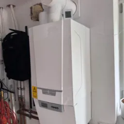 Biały, wiszący piec gazowy De Dietrich z podłączonymi rurami miedzianymi, zamontowany na pomalowanej ścianie w pomieszczeniu technicznym. Widoczny termostat i plecak obok.