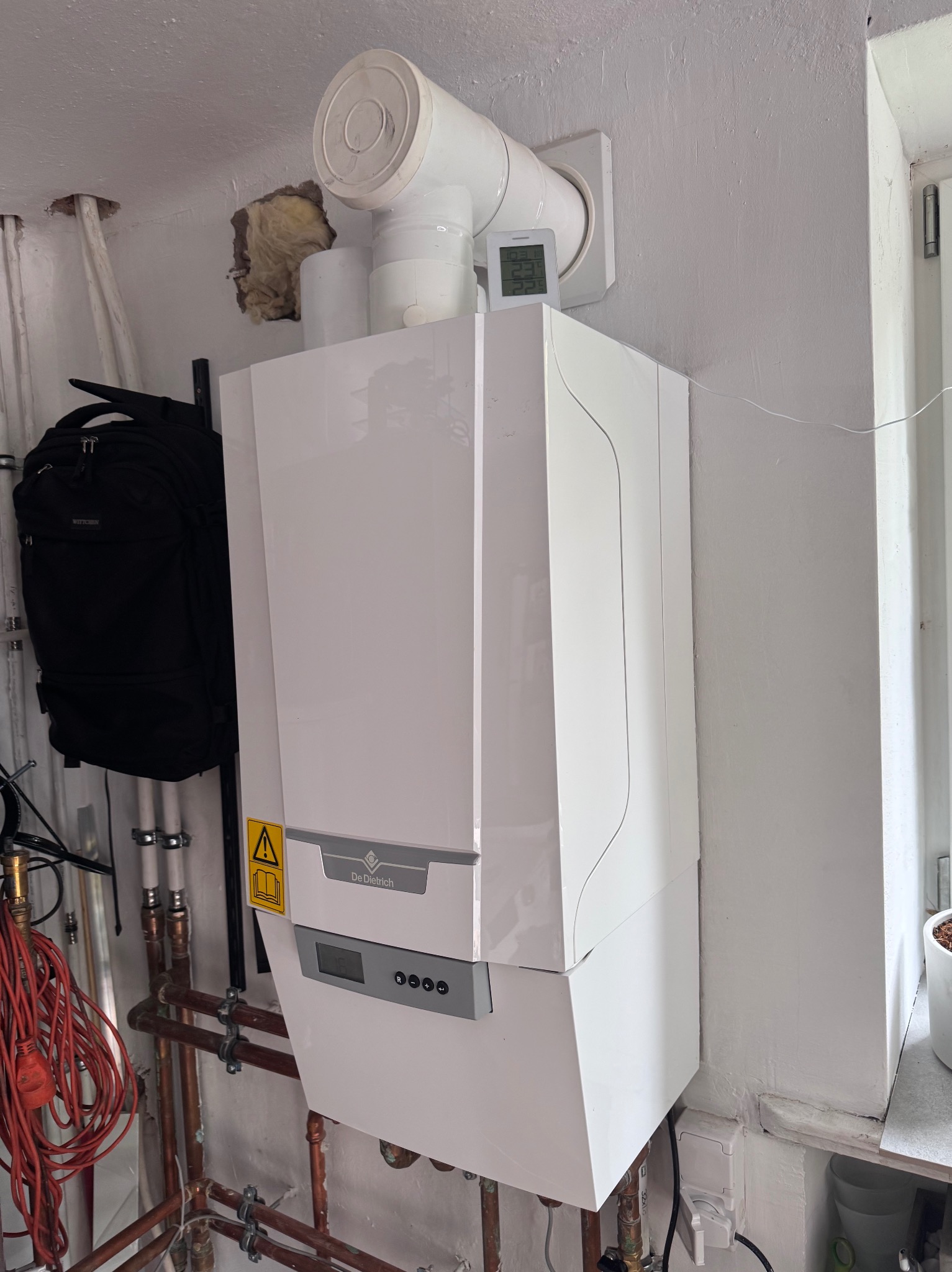 Biały, wiszący piec gazowy De Dietrich z podłączonymi rurami miedzianymi, zamontowany na pomalowanej ścianie w pomieszczeniu technicznym. Widoczny termostat i plecak obok.