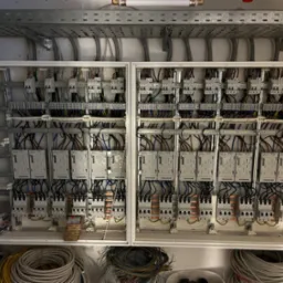 Rozbudowana tablica rozdzielcza w białej obudowie, widoczne liczne bezpieczniki, przewody i szyny montażowe, kable leżące na dole obudowy, szyny kablowe pod sufitem.