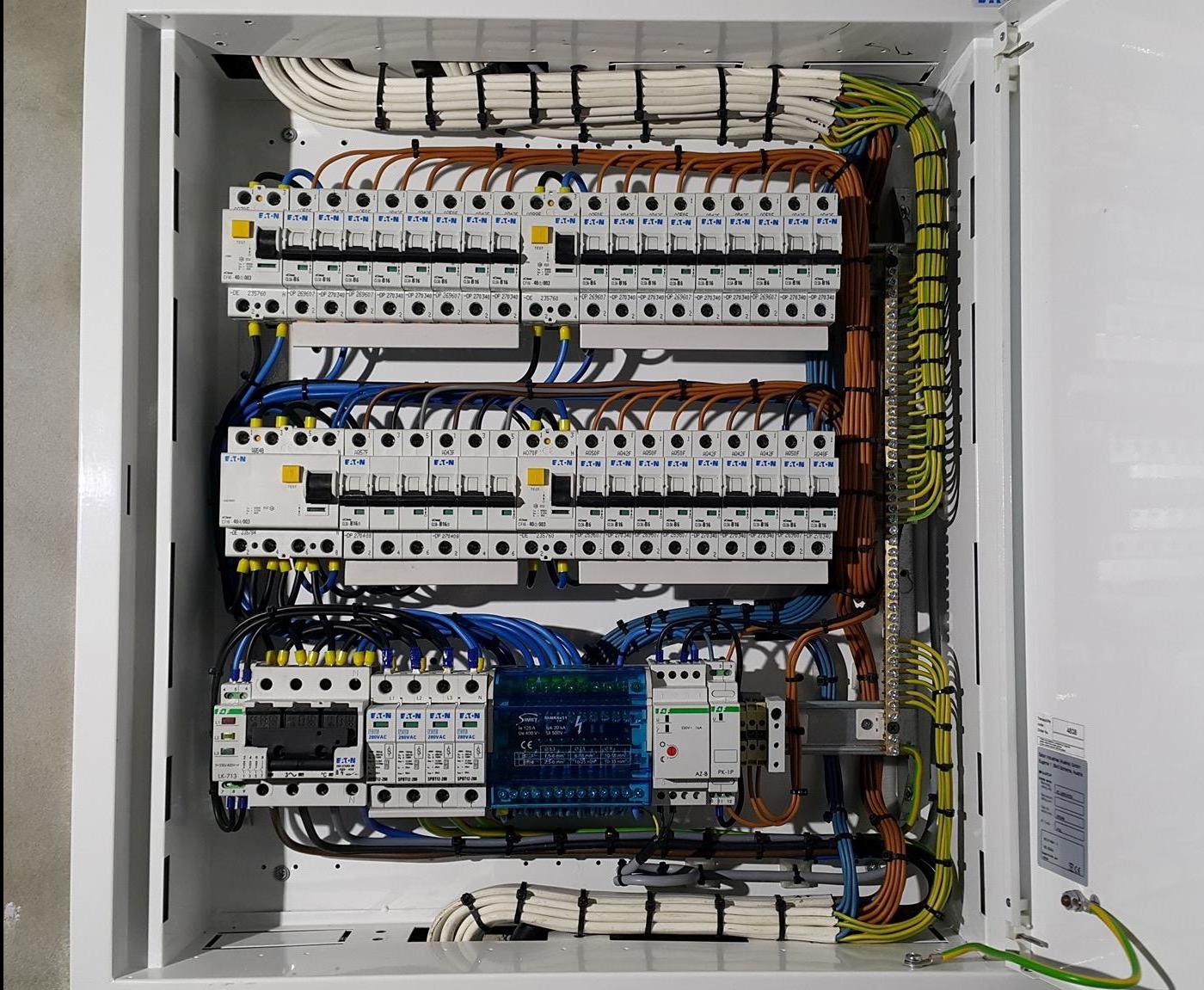 Wewnętrzny widok rozdzielnicy elektrycznej z zabezpieczeniami Eaton, starannie ułożonymi przewodami w różnych kolorach i złączami, zamontowanej w białej obudowie.