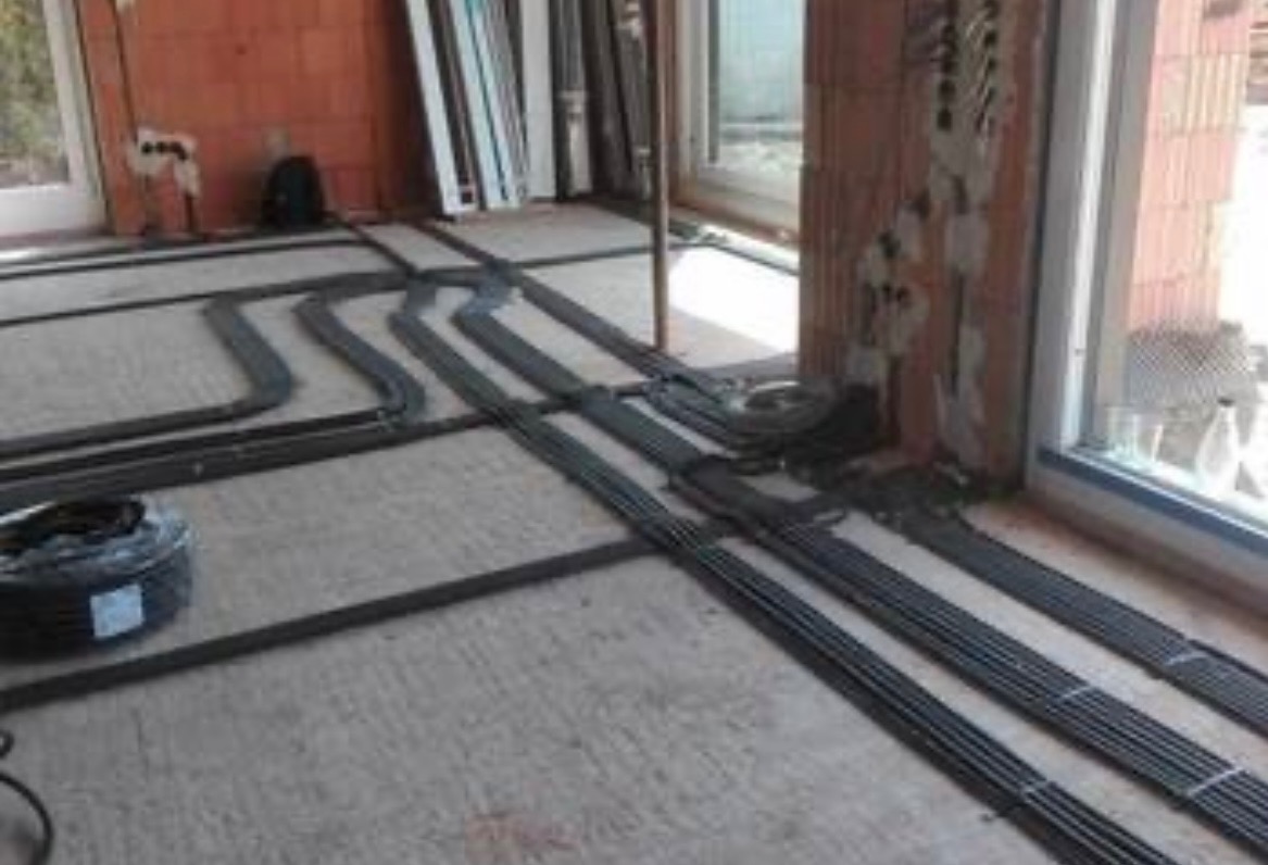 Instalacja ogrzewania podłogowego z rur PEX w surowym stanie, widoczne rozłożone pętle i zwoje rur na betonowej posadzce, ściany z cegły dziurawki, okna PCV.