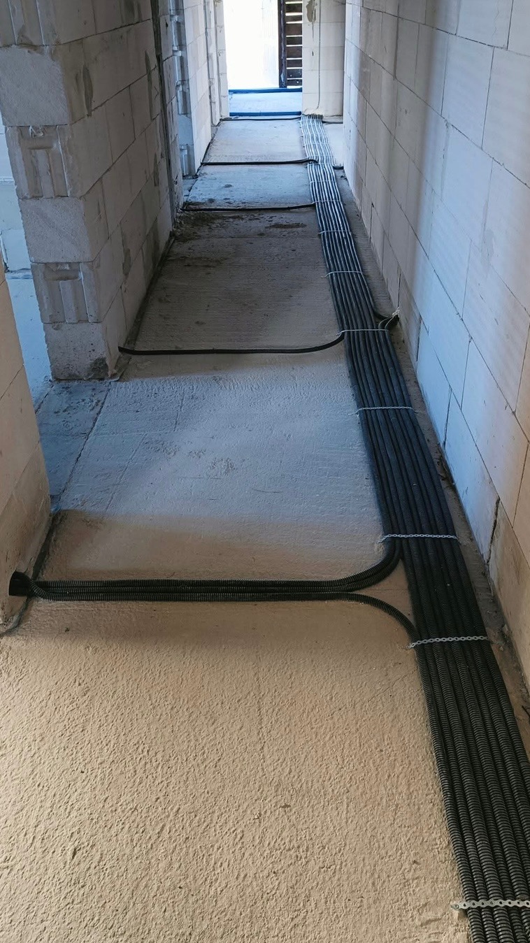 Korytarz w stanie surowym z ułożonymi wzdłuż ściany wiązkami czarnych peszli elektrycznych, przymocowanych metalowymi uchwytami do ściany z betonu komórkowego.
