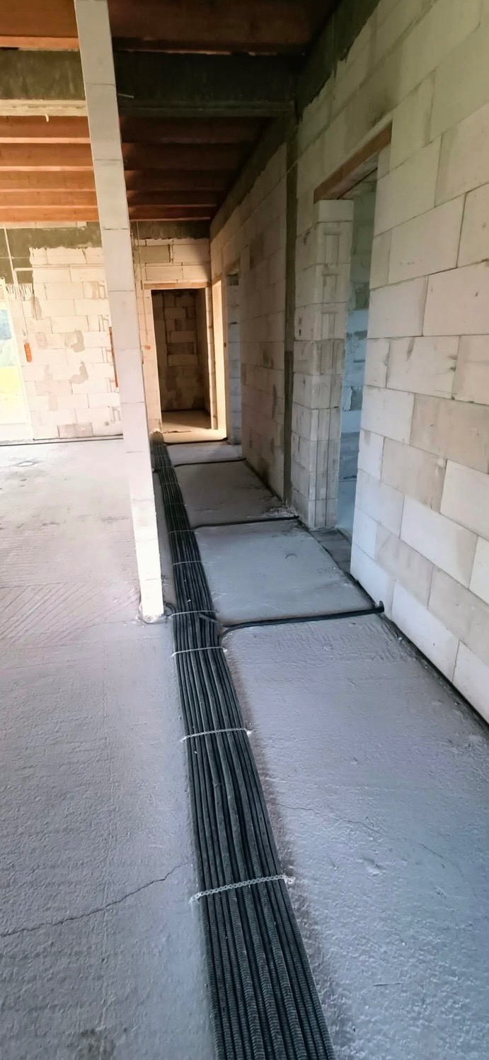 Rury instalacyjne w peszlu ułożone na betonowej podłodze w surowym stanie, widoczne ściany z bloczków i drewniany strop.