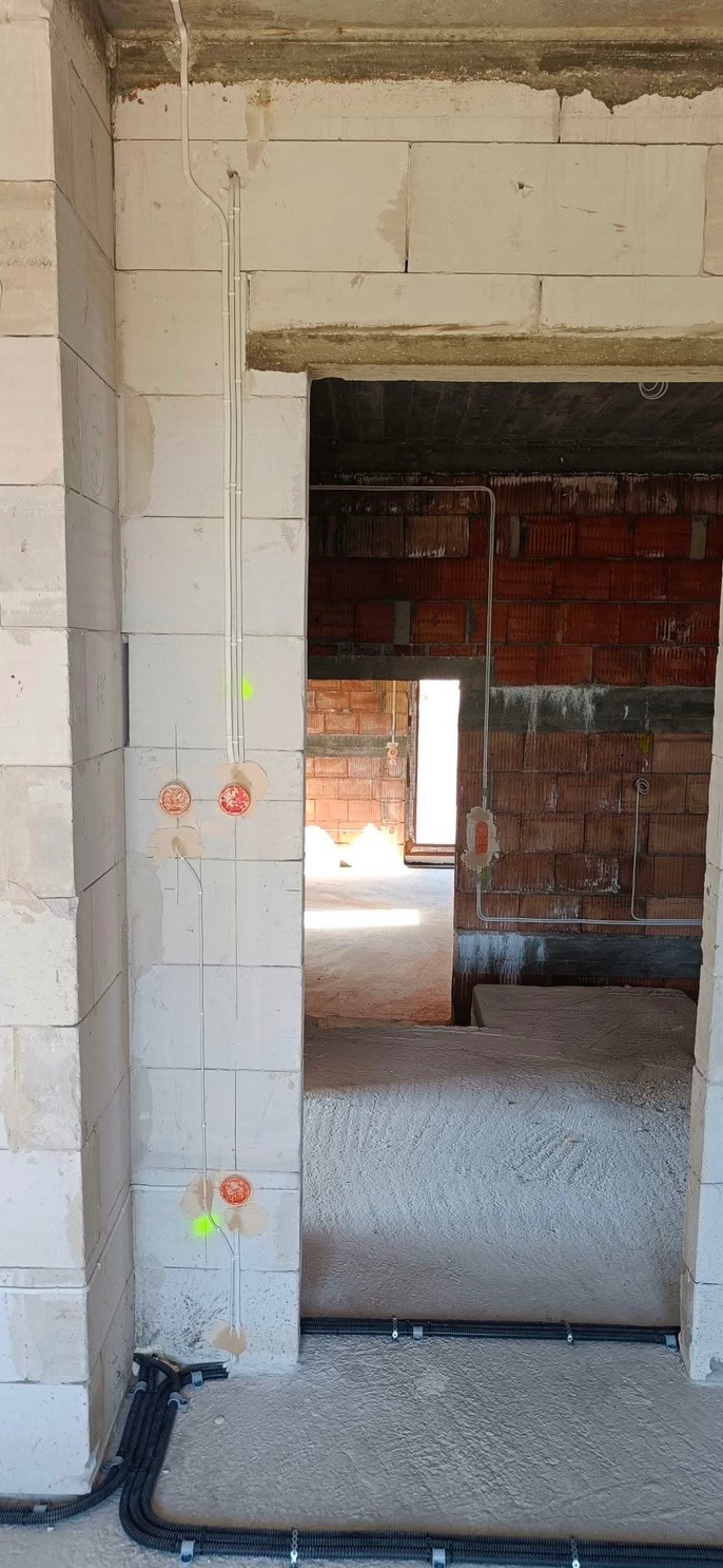 Wewnętrzna instalacja elektryczna w surowym stanie budynku, widoczne kable w peszlach przymocowane do ściany z betonu komórkowego oraz puszki instalacyjne z pomarańczowymi zaślepkami.