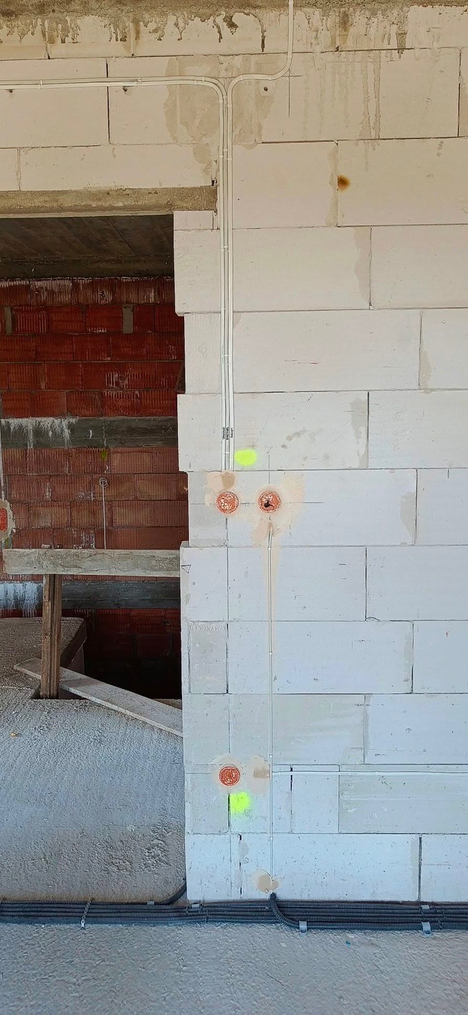 Wewnętrzna instalacja elektryczna w surowym budynku, widoczne kable w peszlach i puszki elektryczne w ścianie z betonu komórkowego, oznaczone fluorescencyjnymi markerami.