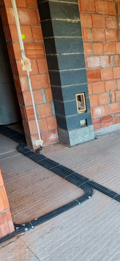 Instalacja elektryczna w peszlach ułożona na podłodze w trakcie budowy, widoczne puszki elektryczne i przewody na ścianie z czerwonej cegły.