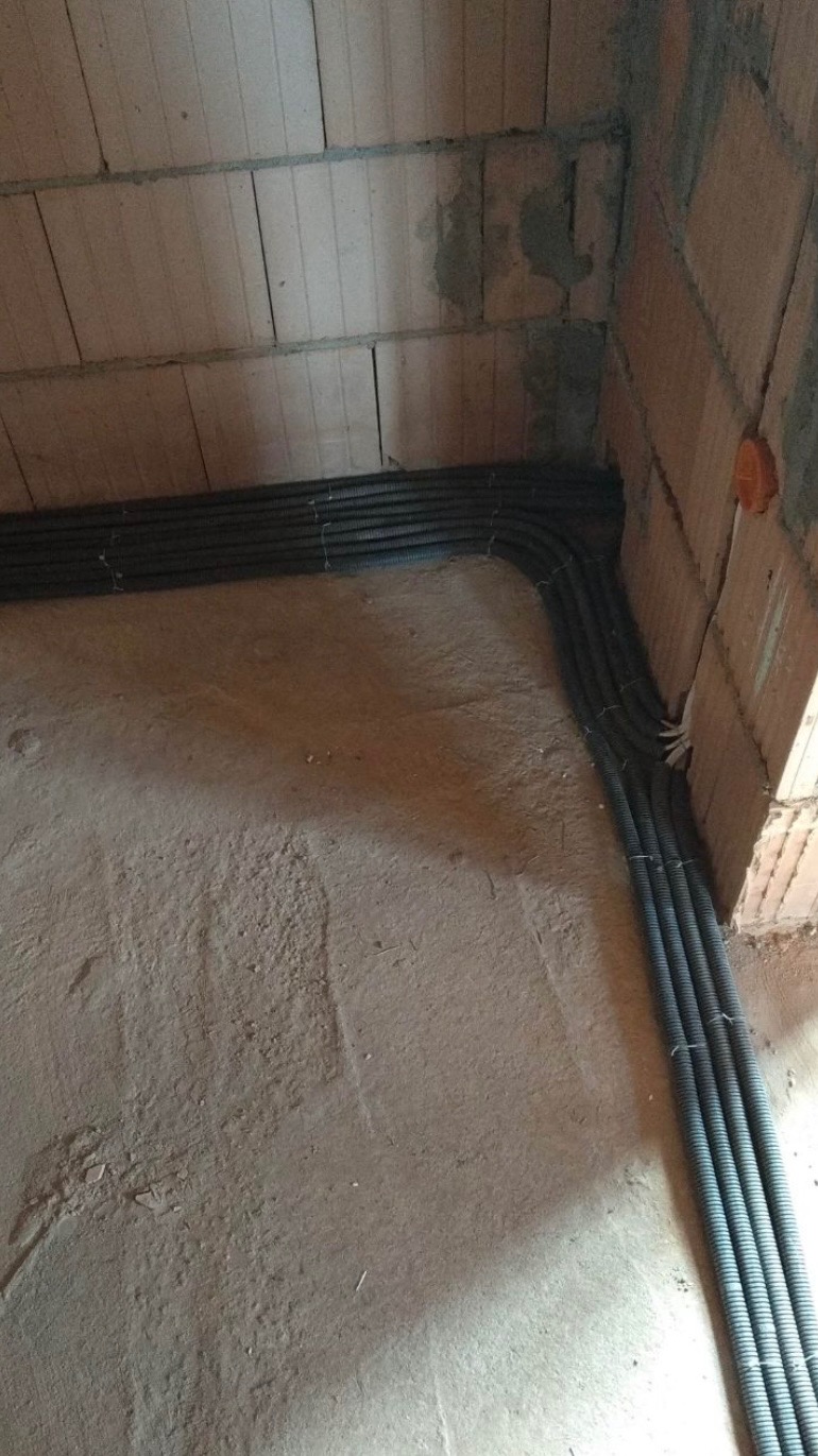 Rury instalacyjne w peszlu ułożone wzdłuż ściany z pustaków ceramicznych i szarej, zacieranej posadzki w surowym stanie.