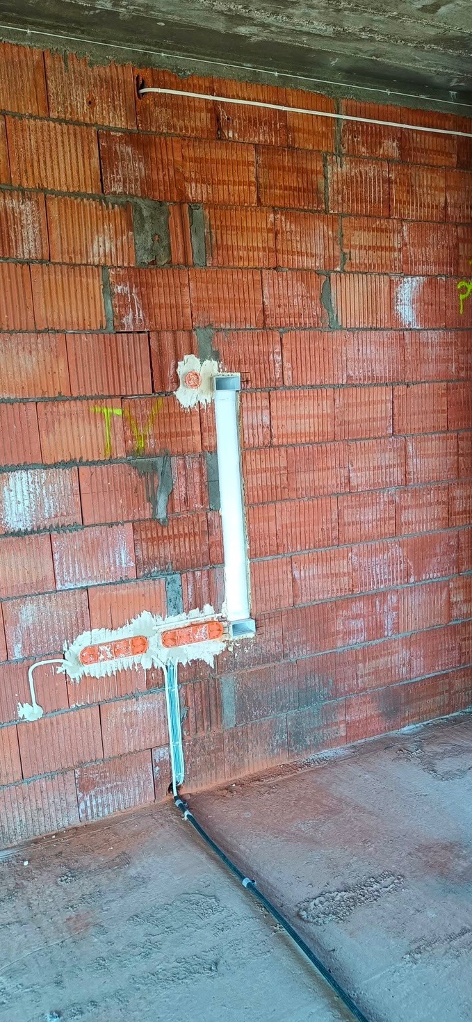 Czerwona ściana z pustaków z widocznymi przewodami elektrycznymi w peszlu i puszkami instalacyjnymi zamontowanymi w ścianie, częściowo zamurowanymi zaprawą.