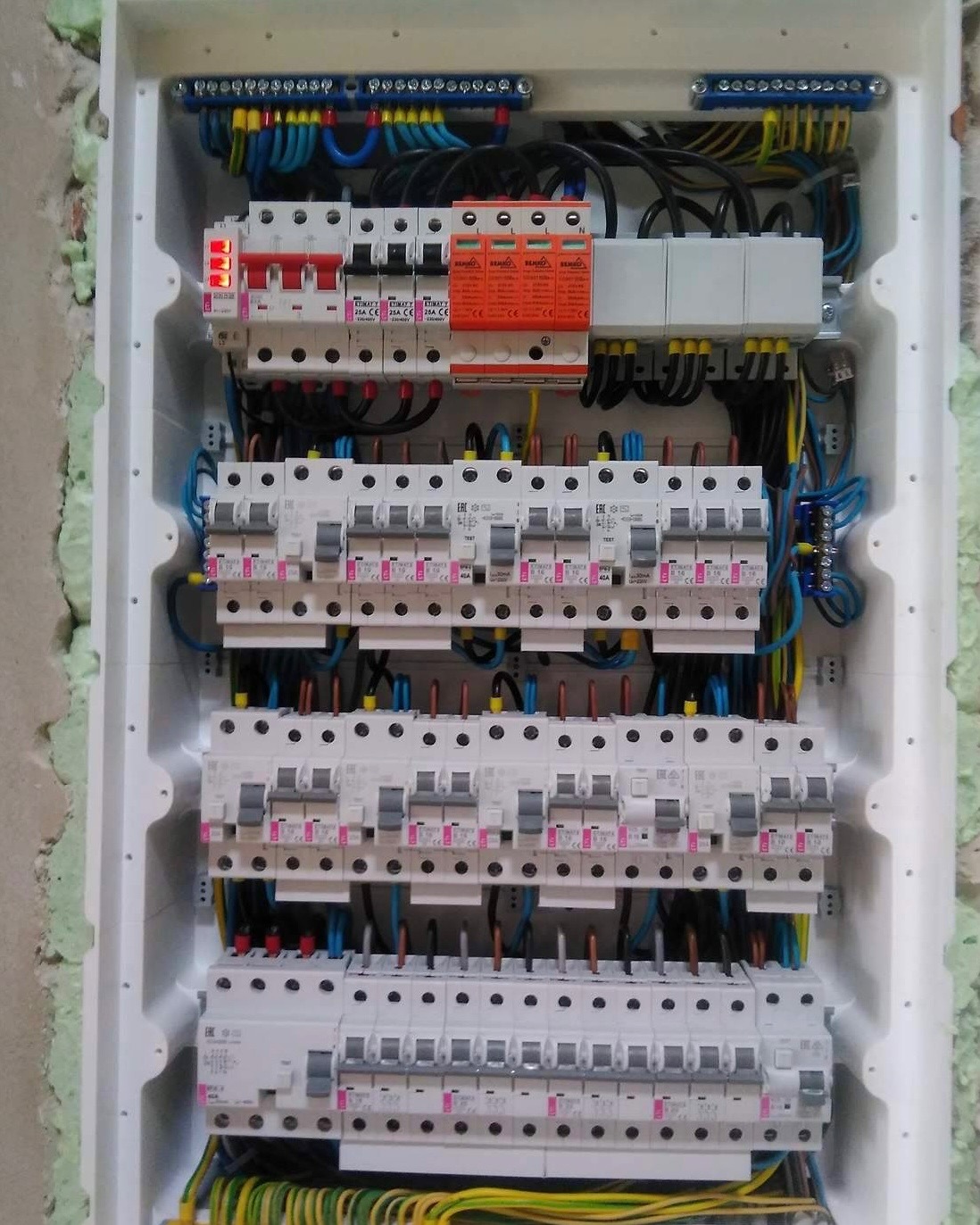 Rozdzielnica elektryczna w białej obudowie z licznymi bezpiecznikami, przewodami w różnych kolorach i zabezpieczeniami przeciwprzepięciowymi, widoczne elementy montażu w ścianie.