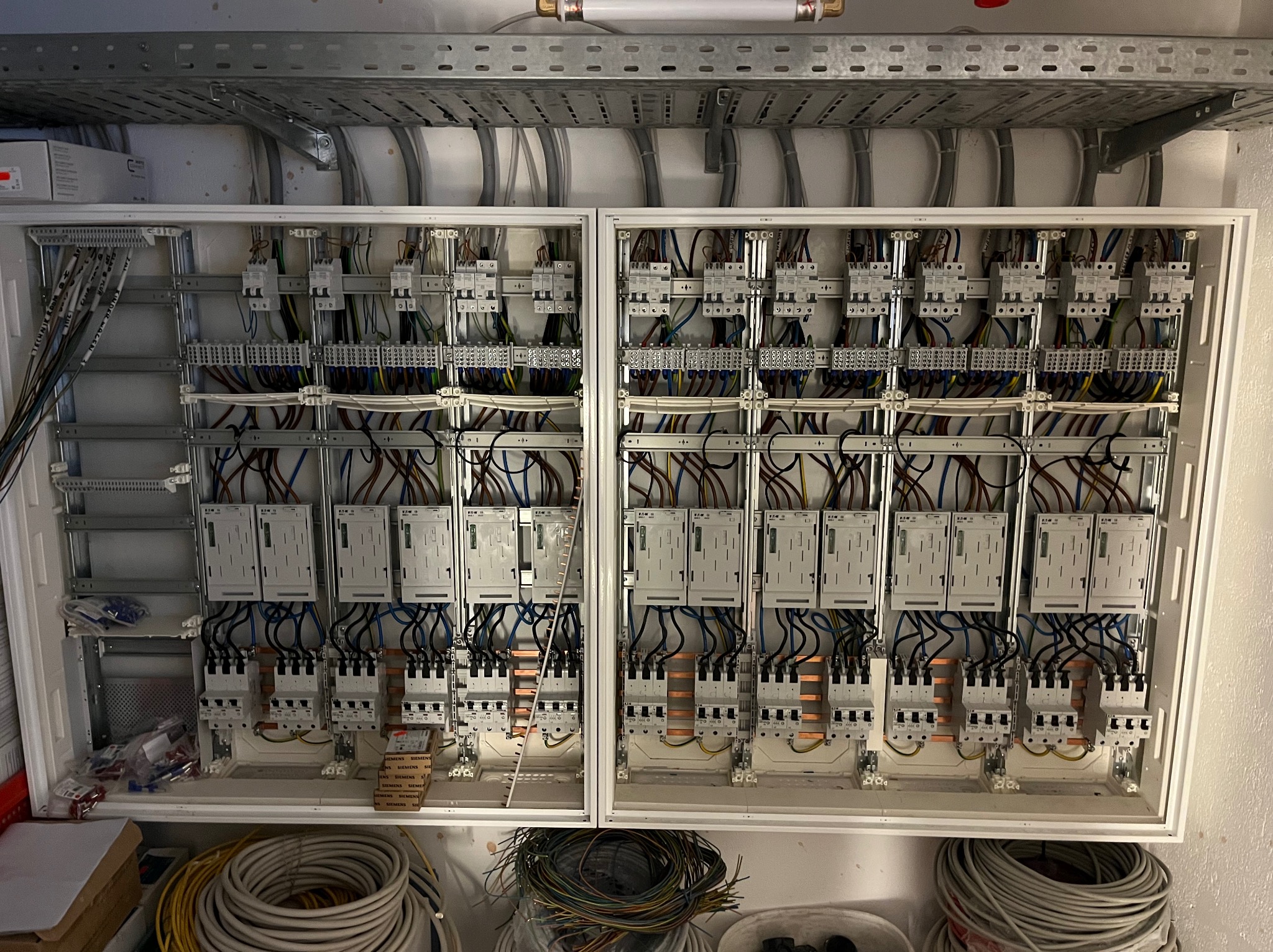 Rozbudowana tablica rozdzielcza w białej obudowie, widoczne liczne bezpieczniki, przewody i szyny montażowe, kable leżące na dole obudowy, szyny kablowe pod sufitem.