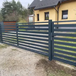 Nowoczesna, pozioma brama wjazdowa w kolorze grafitowym, z widocznym zamkiem, na tle domu z żółtą elewacją i szarą dachówką. Minimalistyczny design.