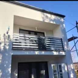 Nowoczesna balustrada balkonowa z poziomych profili, zamontowana w domu w Cieszynie. Widoczne rusztowanie, błękitne niebo i cień drzewa na elewacji.