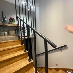 DANMAR Marcin Małyjurek - Nowoczesne schody drewniane z czarną, metalową balustradą. Widoczne stopnie z jasnego drewna i minimalistyczny design balustrady z pionowych prętów.