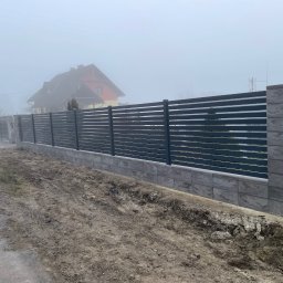 DANMAR Marcin Małyjurek - Nowoczesne ogrodzenie panelowe z podmurówką z szarej kostki, widoczne w mglisty dzień. W tle dom z czerwonym dachem.
