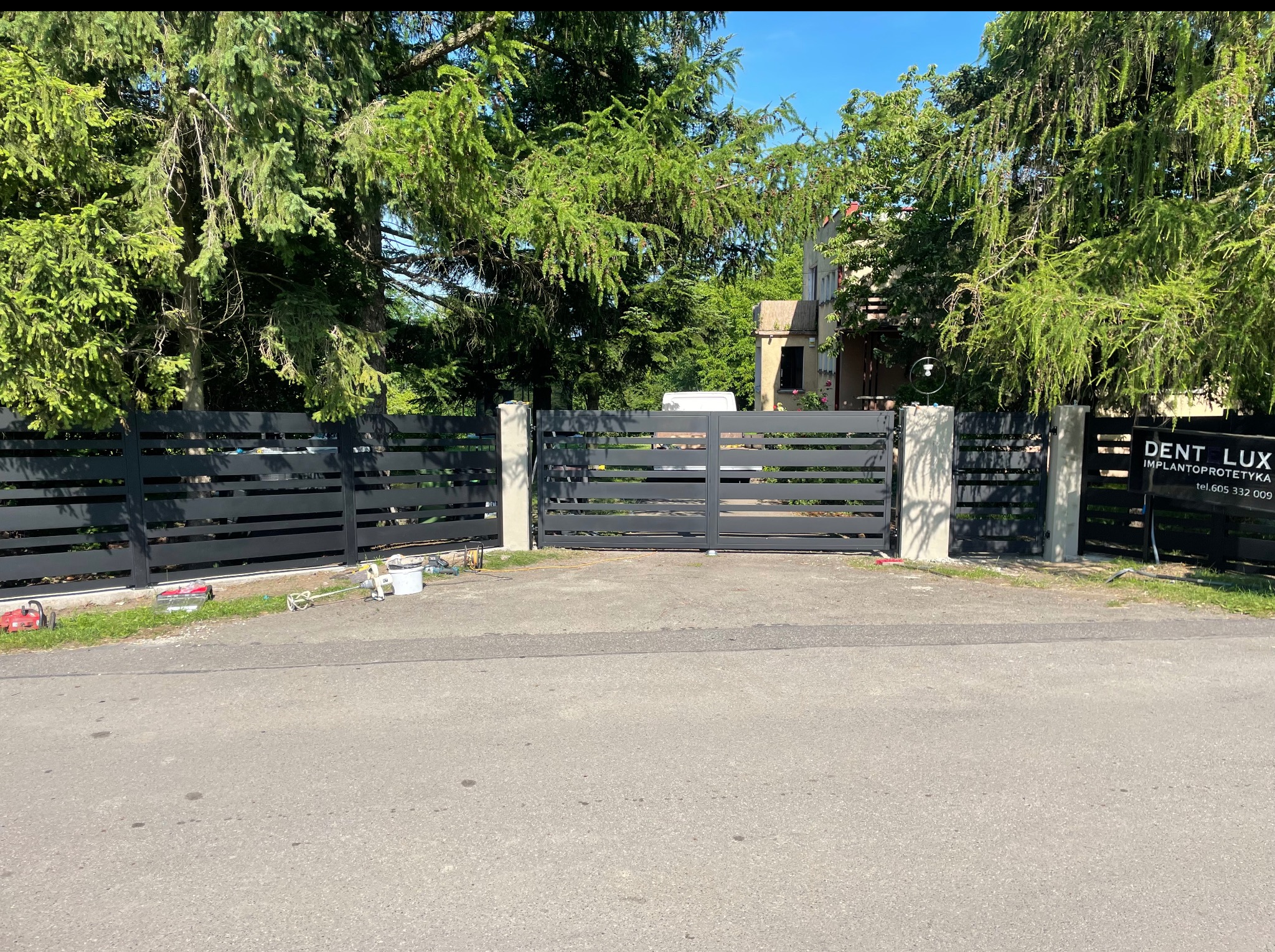 Nowoczesne, poziome ogrodzenie panelowe w kolorze antracytowym z betonowymi słupkami. Widoczna brama wjazdowa i narzędzia montażowe. Tło: zieleń drzew.