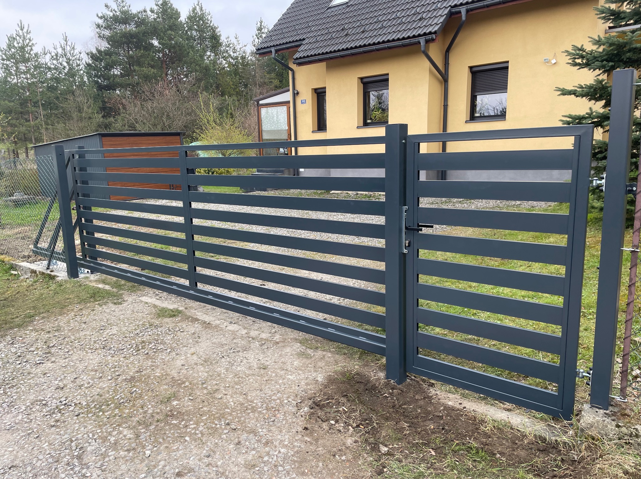 Nowoczesna, pozioma brama wjazdowa w kolorze grafitowym, z widocznym zamkiem, na tle domu z żółtą elewacją i szarą dachówką. Minimalistyczny design.