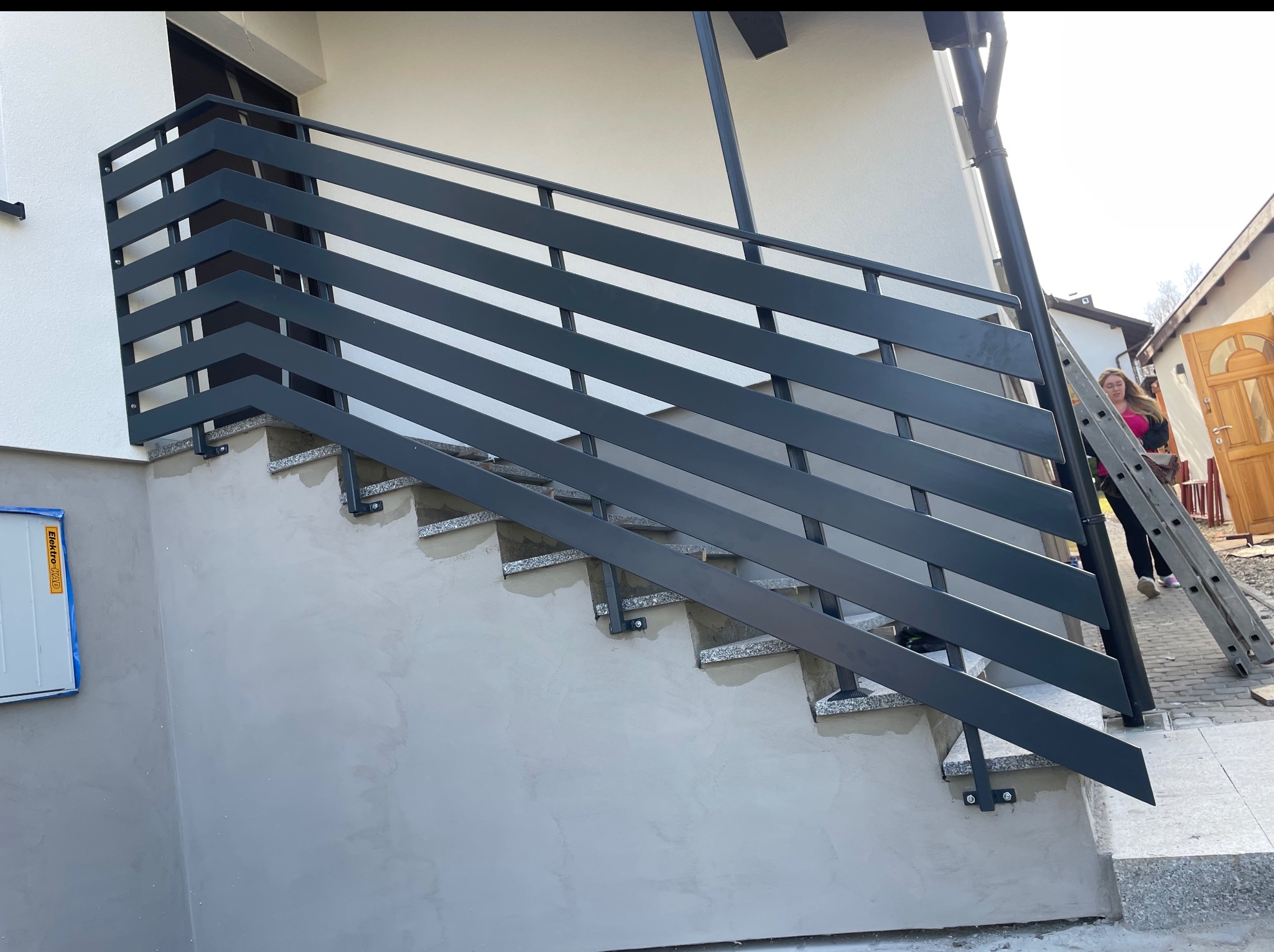 Nowoczesna, metalowa balustrada schodowa w kolorze grafitowym, zamontowana przy wejściu do budynku. Widoczne granitowe stopnie i fragment elewacji.