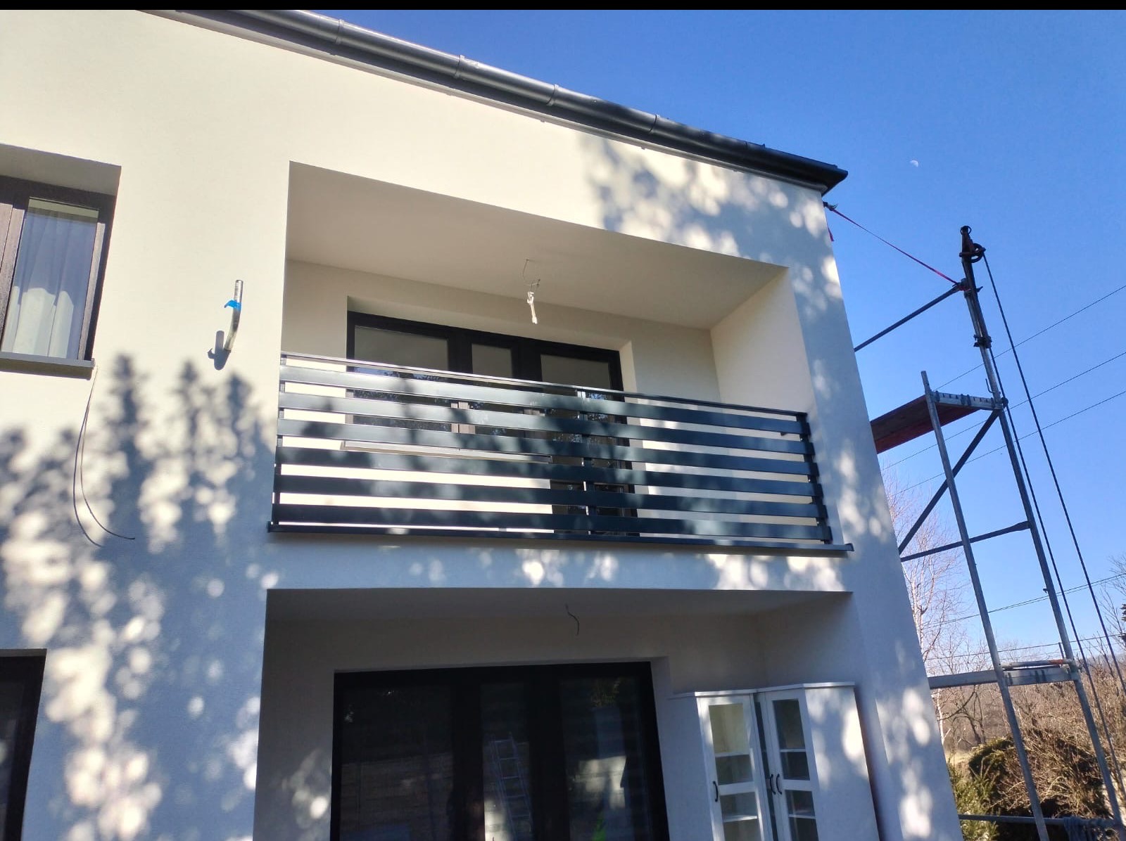 Nowoczesna balustrada balkonowa z poziomych profili, zamontowana w domu w Cieszynie. Widoczne rusztowanie, błękitne niebo i cień drzewa na elewacji.