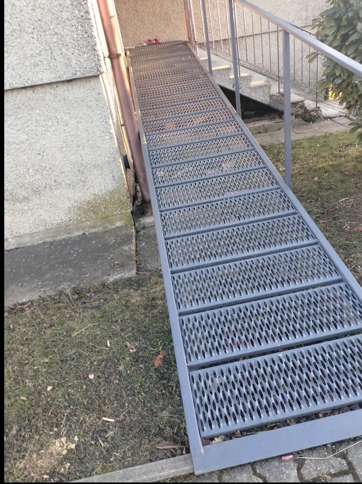Metalowa rampa z perforowaną powierzchnią prowadząca do schodów, zainstalowana przy budynku z szarą elewacją. Widoczny fragment trawnika i rynna.
