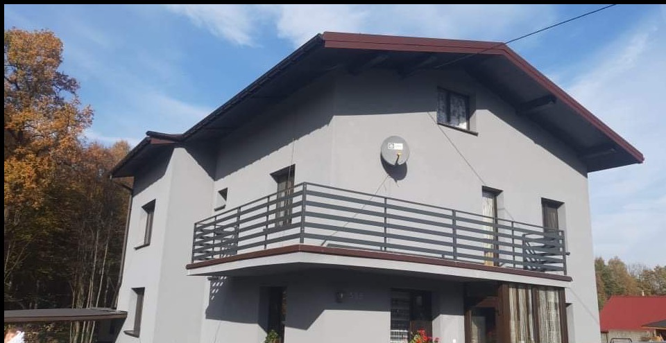 Szary dom z balkonem i metalową balustradą na tle jesiennych drzew i błękitnego nieba. Widoczna antena satelitarna.