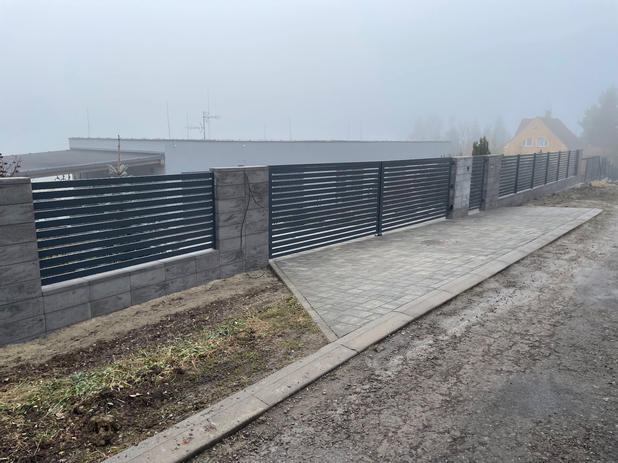 Nowoczesne ogrodzenie panelowe z szarej kostki i poziomych przęseł, widoczne w pochmurny dzień. Minimalistyczny design, świeżo ułożona kostka brukowa.