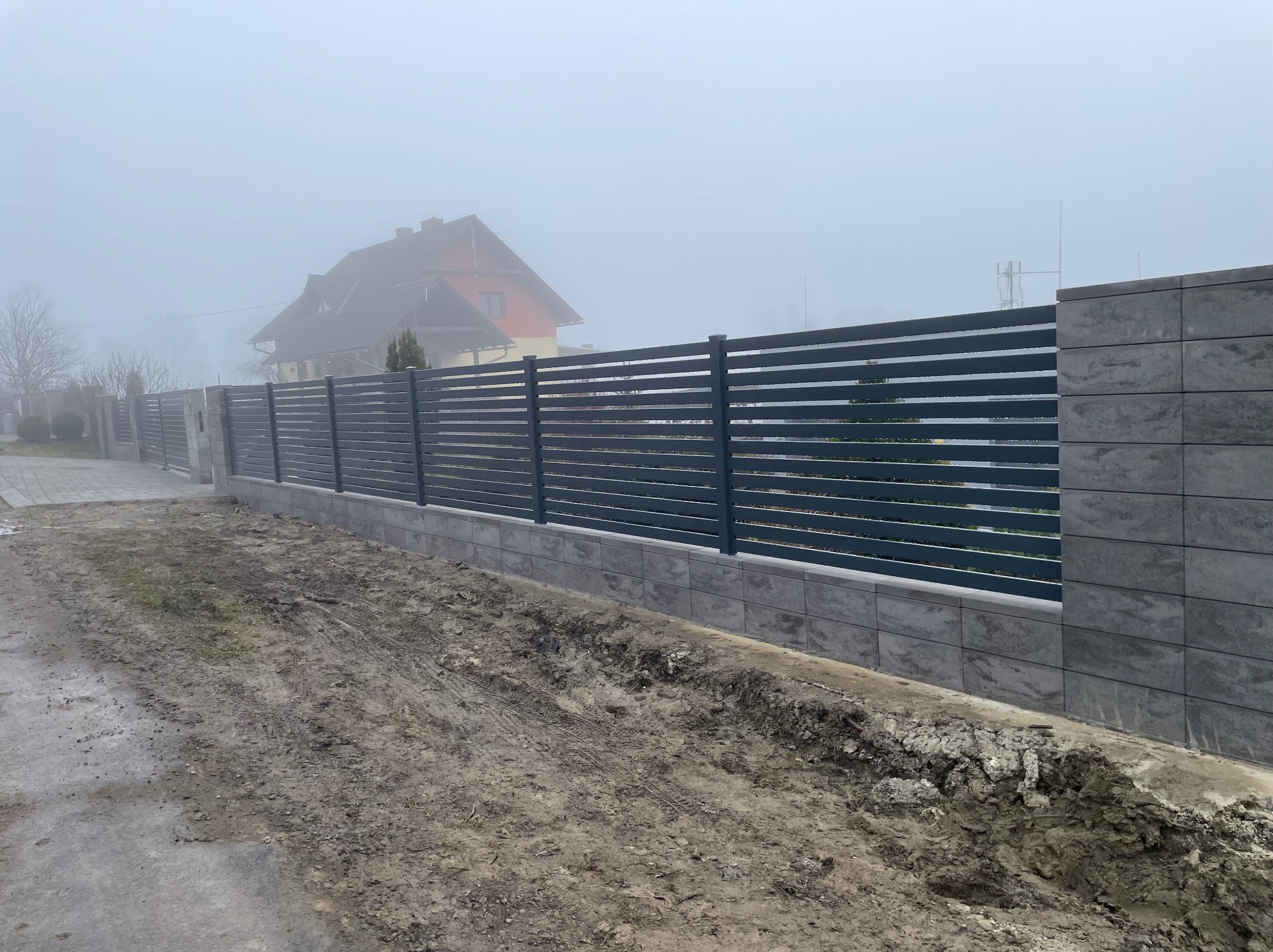 Nowoczesne ogrodzenie panelowe z podmurówką z szarej kostki, widoczne w mglisty dzień. W tle dom z czerwonym dachem.