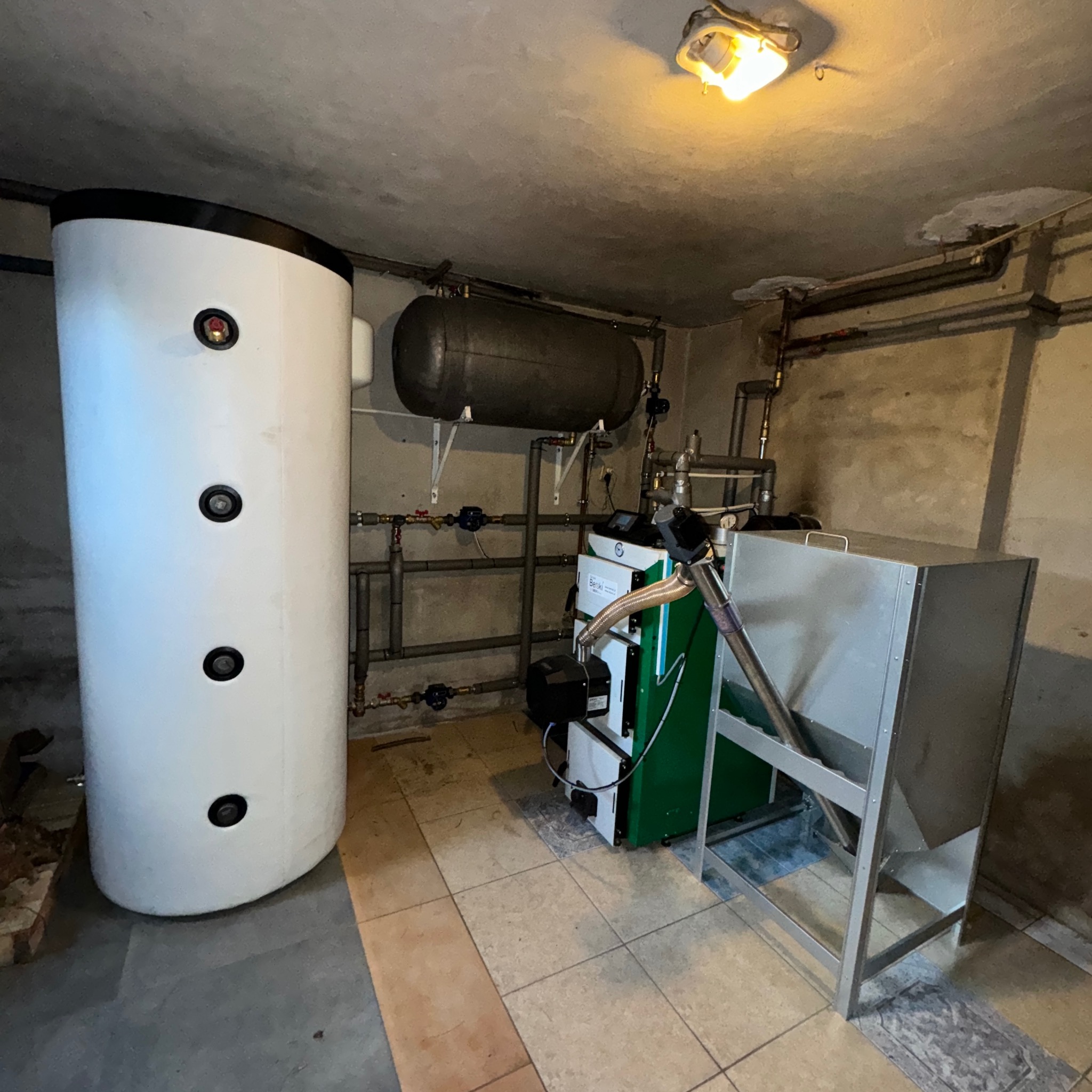Zainstalowany piec na paliwo stałe z podajnikiem i zbiornikiem buforowym w kotłowni. Widoczne rury instalacji grzewczej i oświetlenie. Ujęcie z niskiego kąta.