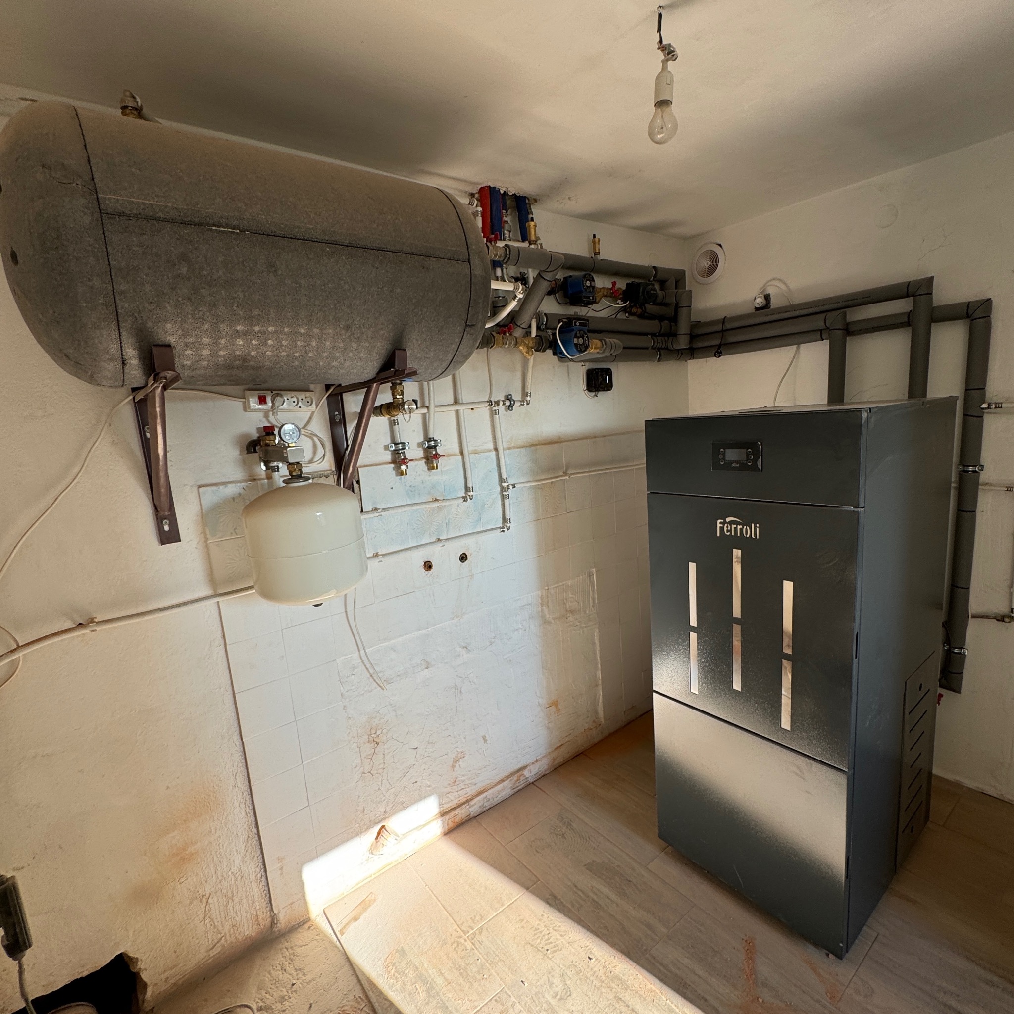 Szary piec Ferroli na paliwo stałe w kotłowni z instalacją CO i zbiornikiem akumulacyjnym. Widoczne rury, zawory i manometry. Oświetlenie żarówką.