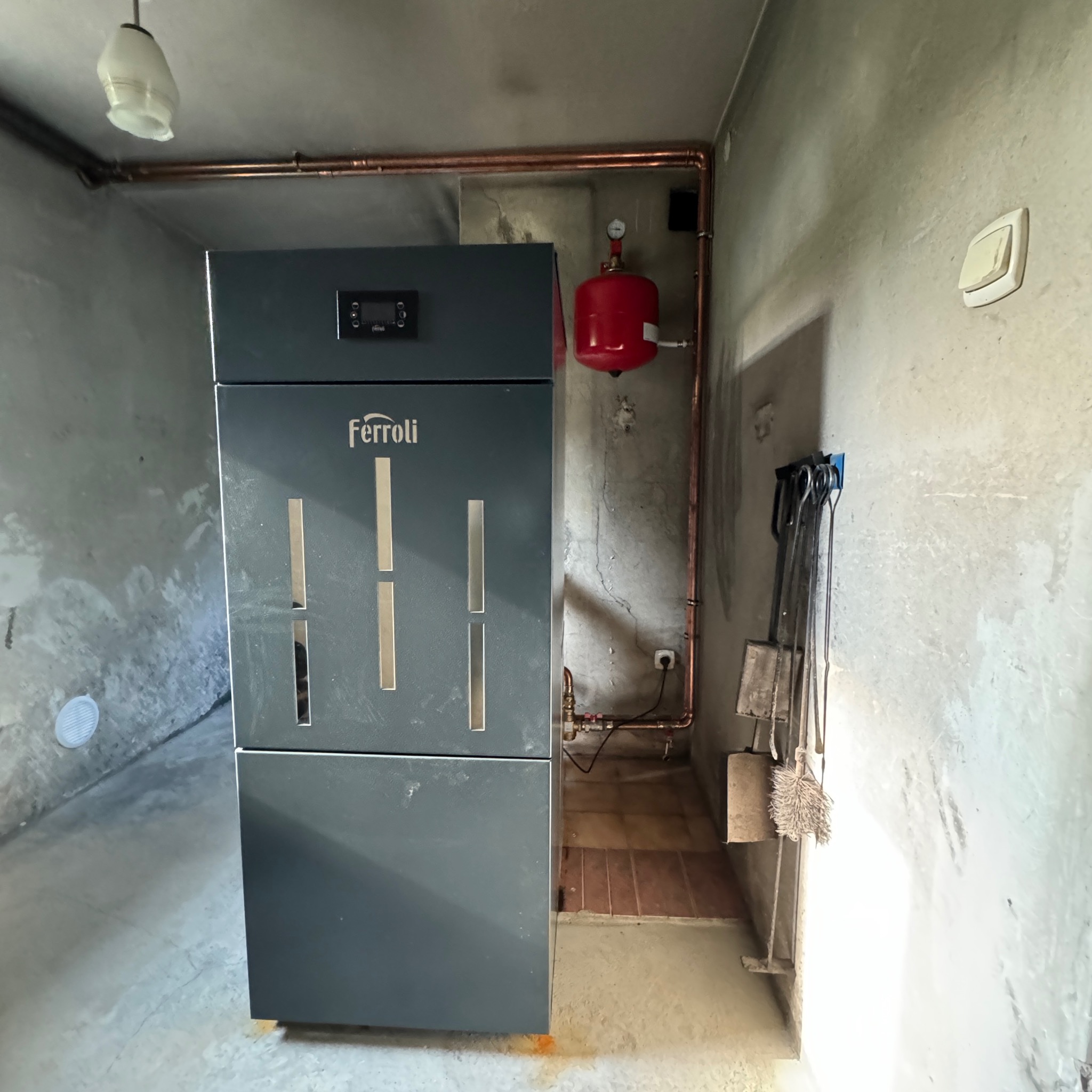 Szary piec Ferroli na paliwo stałe w kotłowni z instalacją miedzianą, naczyniem wzbiorczym i zestawem narzędzi kominkowych na tle surowych ścian.