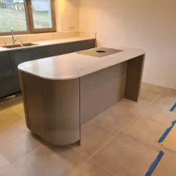 Beżowa wyspa kuchenna z wbudowaną płytą grzewczą na tle okna i zabudowy kuchennej w szarym odcieniu, widoczne lampy wiszące i fragment podłogi z zabezpieczeniami.