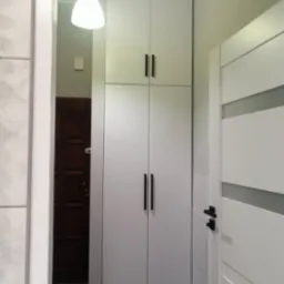 Biała szafa do sufitu z czarnymi, podłużnymi uchwytami, ustawiona w przedpokoju obok białych drzwi z szarym paskiem, oświetlona wiszącą lampą z białym kloszem.