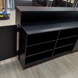 Czarna lada sklepowa z otwartymi drzwiczkami szafki, widoczne półki, obok chłodziarka ekspozycyjna, na podłodze płytki imitujące drewno, w tle narzędzia.