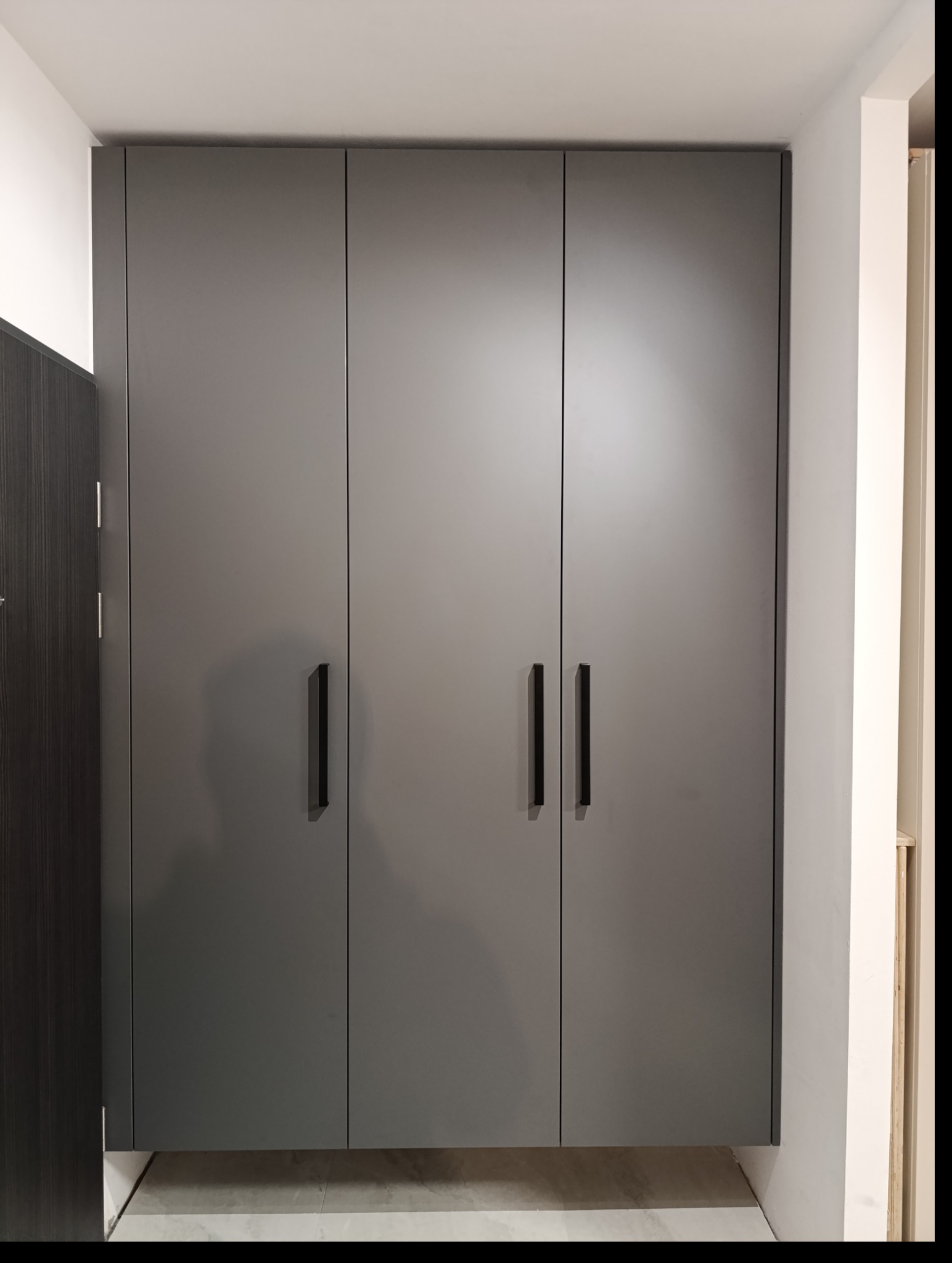 Szara szafa na wymiar z trzema drzwiami i czarnymi, pionowymi uchwytami, zamontowana w białym wnętrzu, z widocznym fragmentem podłogi z szarych płytek.