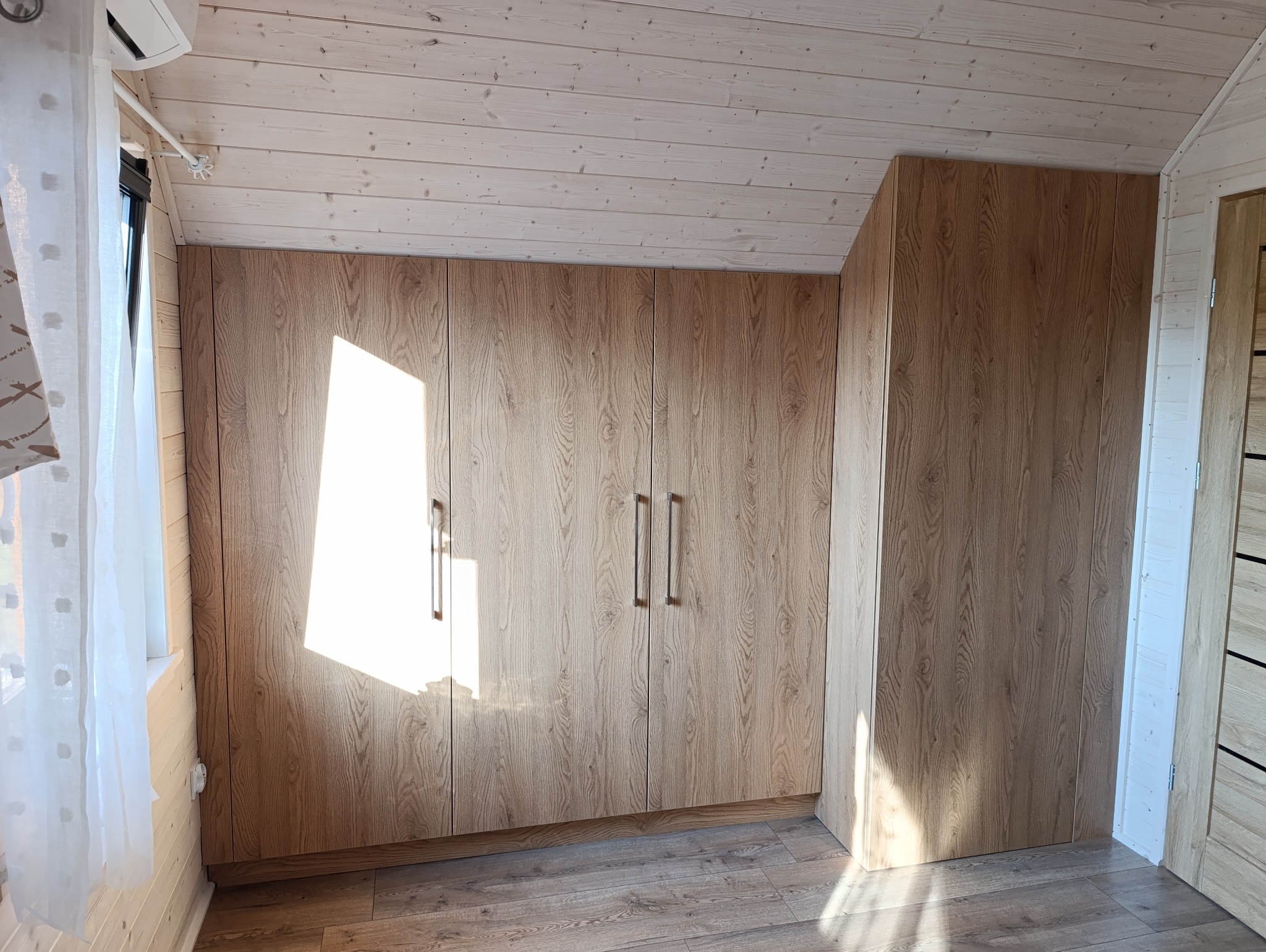 Drewniana szafa z czterema drzwiami, z długimi, metalowymi uchwytami, w jasnym pomieszczeniu z drewnianą podłogą i sufitem, oświetlona padającym światłem słonecznym.