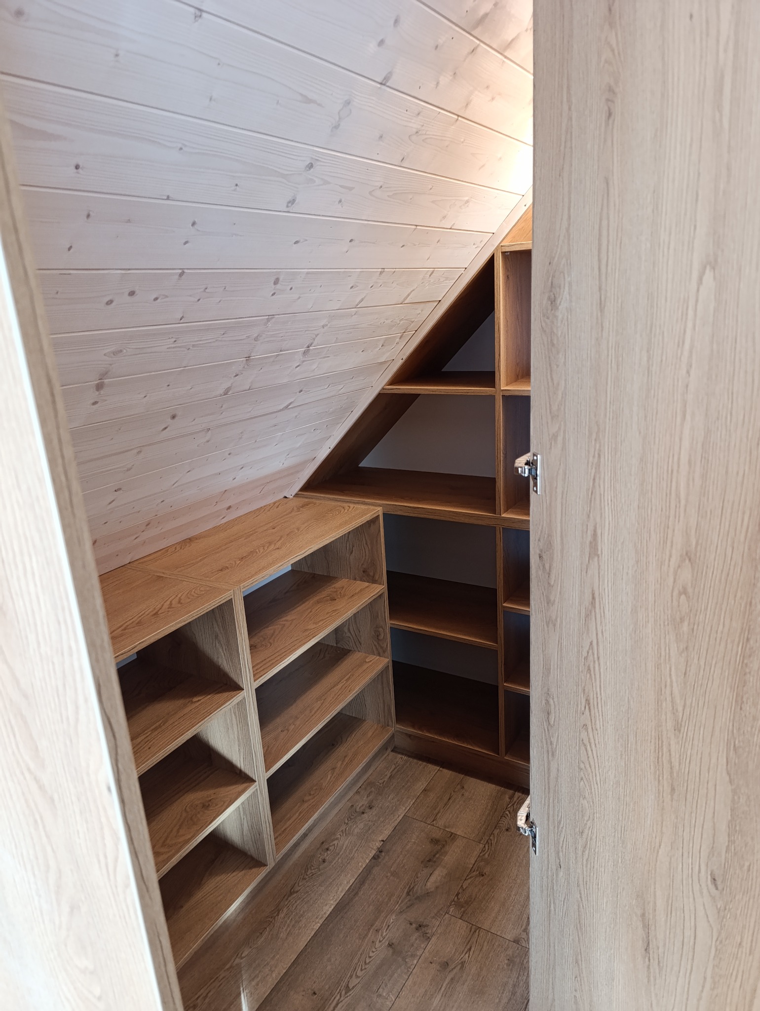 Drewniana zabudowa wnęki na poddaszu ze skośnym sufitem, półki z jasnego drewna w odcieniach brązu.