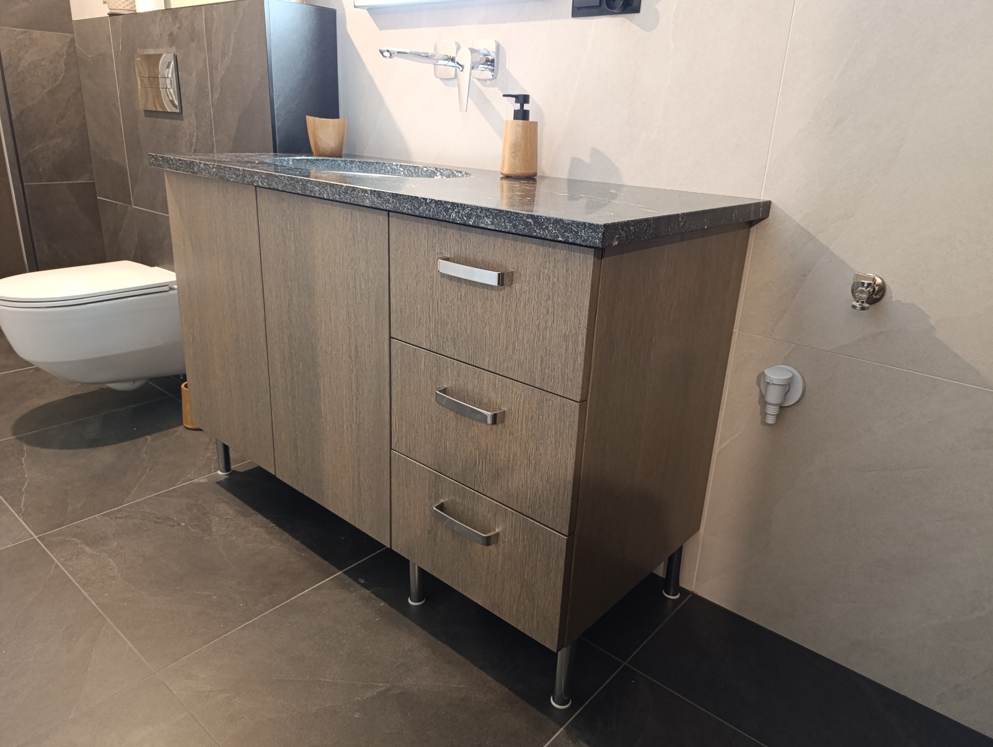 Łazienka z drewnianą szafką pod umywalkę z trzema szufladami, czarnym blatem i chromowanymi uchwytami, widok na toaletę i armaturę.