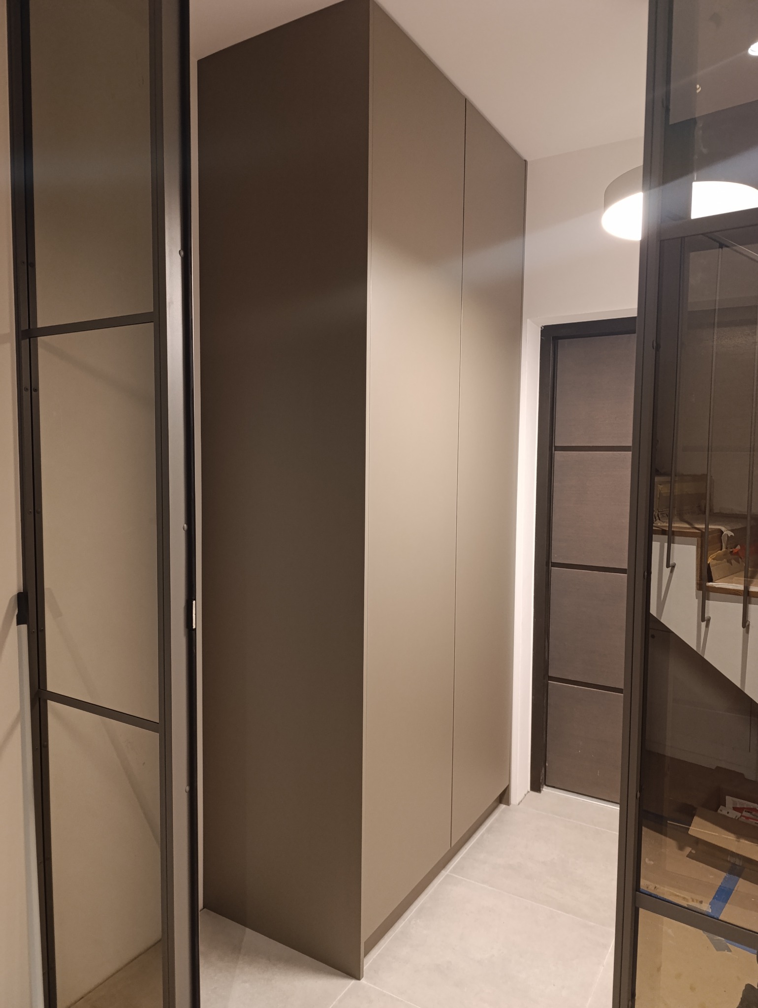 Wysoka, zabudowana szafa w odcieniach beżu, zintegrowana z wnętrzem przedpokoju, widoczna przez szklane drzwi w metalowej ramie.