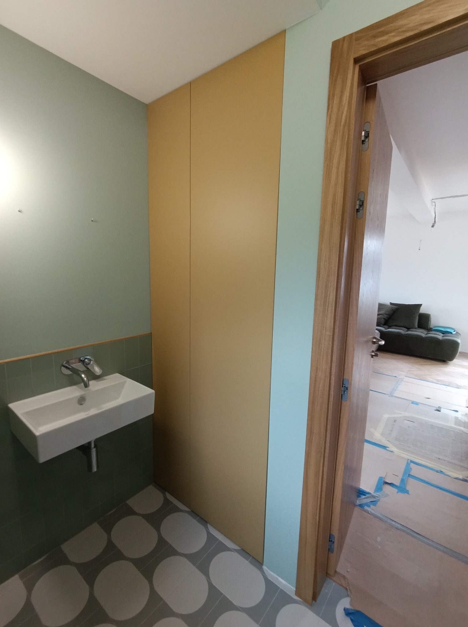 Wnętrze łazienki w trakcie remontu: widoczna zabudowa szafy w kolorze beżowym, umywalka na tle zielonej ściany i drewniana rama drzwi prowadząca do salonu z zabezpieczoną podłogą.