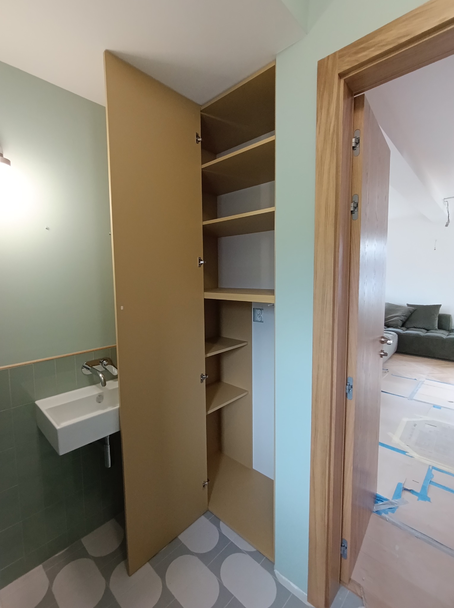 Otwarta szafa z półkami w łazience, widoczna biała umywalka i zielone płytki, z boku drewniana ościeżnica drzwi, w tle salon w trakcie remontu.