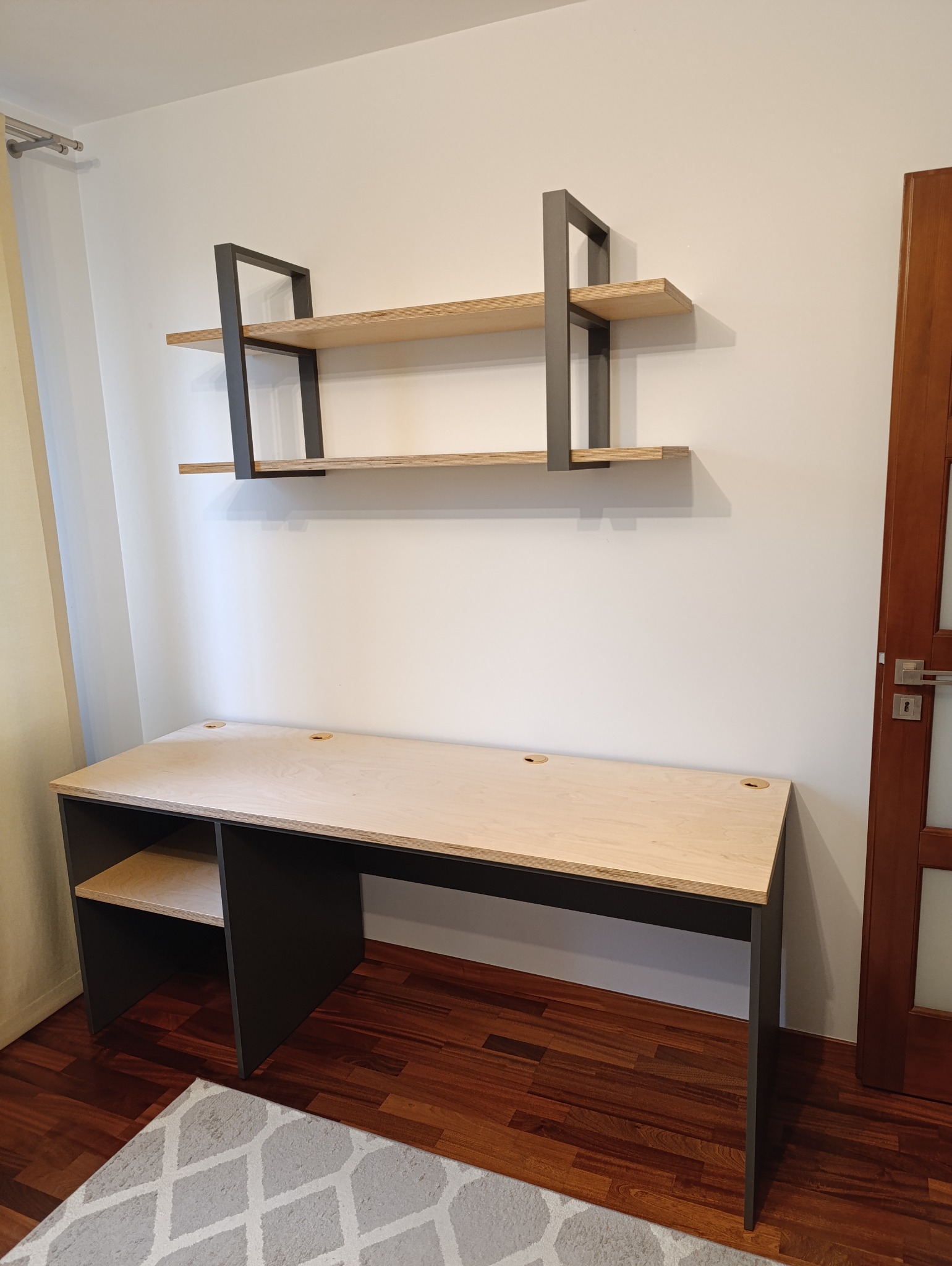 Nowoczesne biurko z jasnego drewna z czarnymi nogami i półką, wisząca półka z dwoma poziomami z drewnianymi blatami i metalową ramą, na tle białej ściany i drewnianej podłogi.