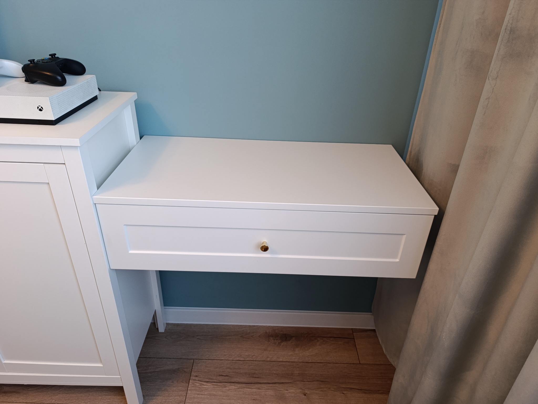 Biała szafka nocna z szufladą i złotym uchwytem, obok konsola do gier na szafce, na tle błękitnej ściany i zasłony.