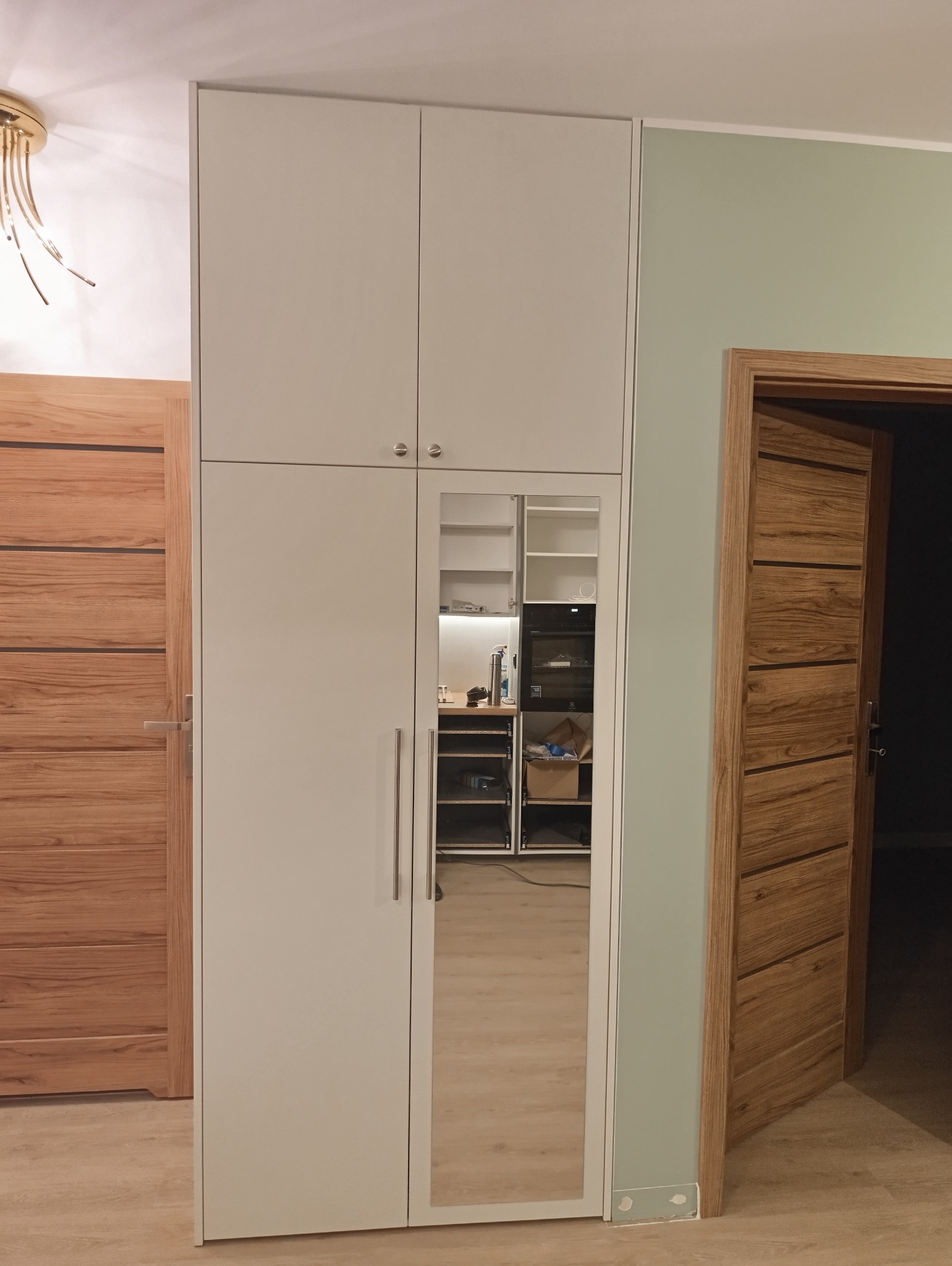 Biała szafa z lustrem wbudowanym w drzwi, widoczne wnętrze z półkami i szufladami, obok drewniane drzwi wejściowe.