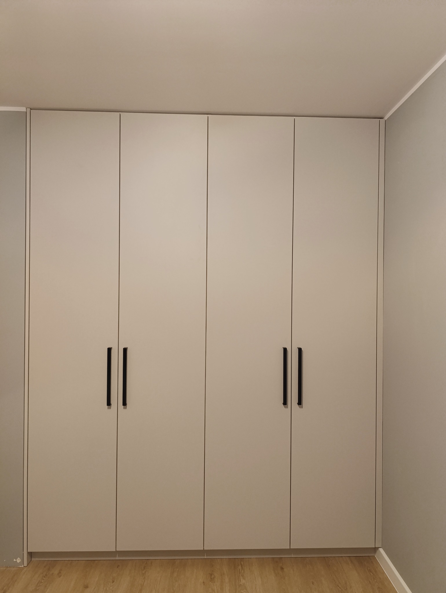 Jasnoszara szafa wnękowa z minimalistycznymi, czarnymi uchwytami, zrealizowana w Kętach. Widoczna podłoga imitująca drewno i fragment szarej ściany.