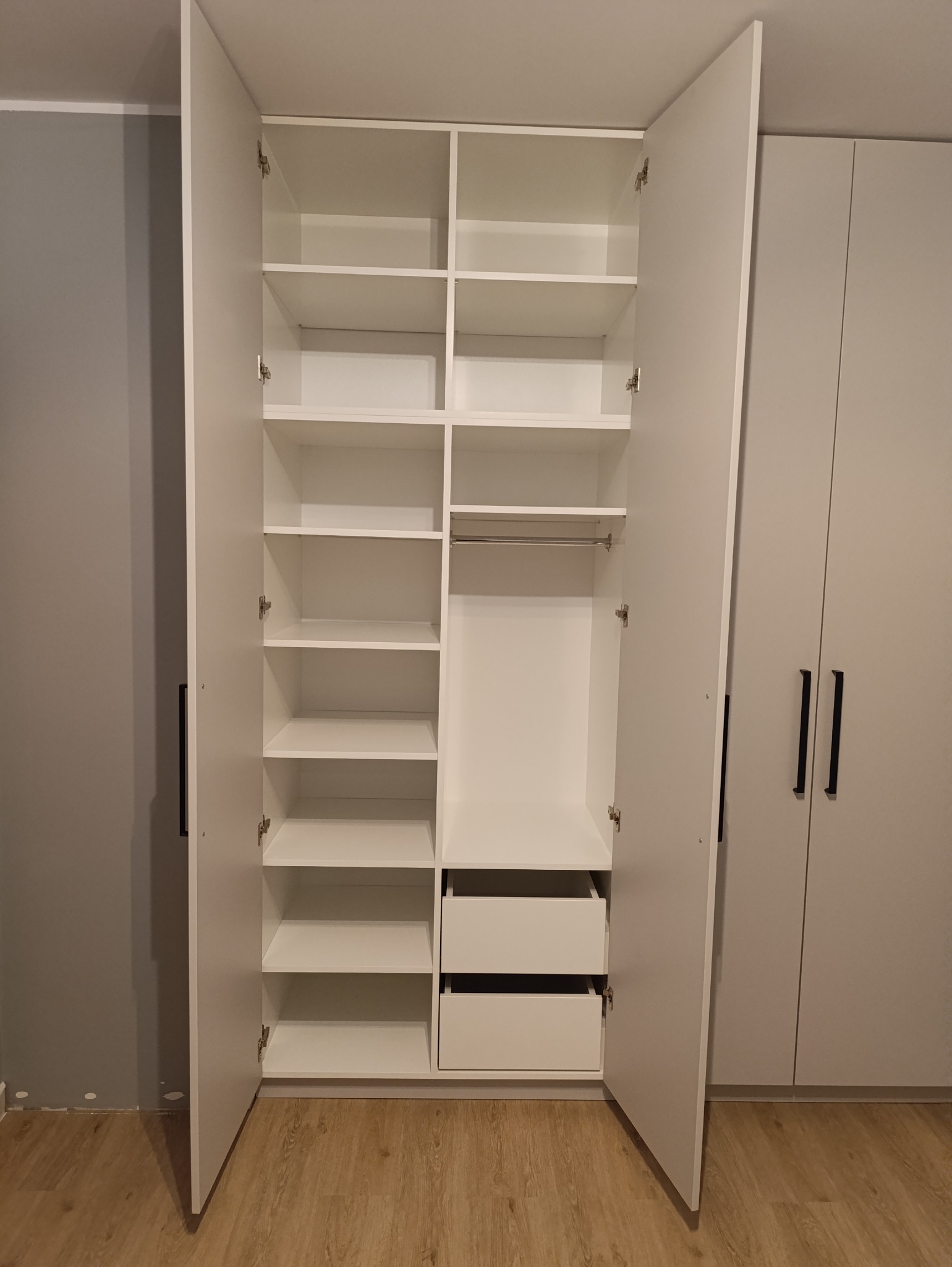 Wnętrze białej, otwartej szafy z półkami, drążkiem i dwoma szufladami, widok na sąsiednią szafę z czarnymi uchwytami.